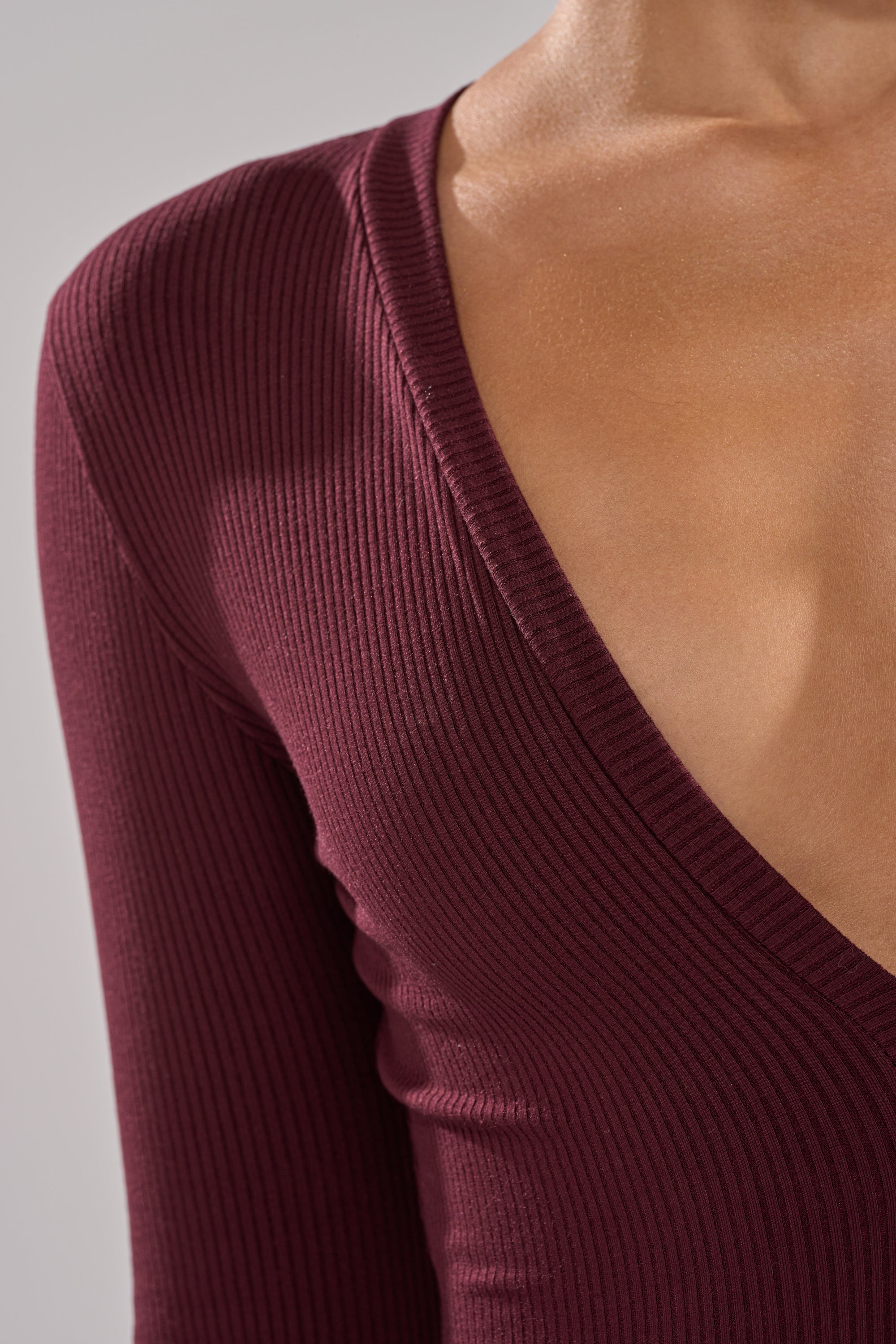 A close-up of a person wearing the DEEP V LONG SLEEVE JUMPSUIT IN RAISIN, featuring a ribbed knit fabric and deep V-neck that accentuates the collarbone and upper chest. 3