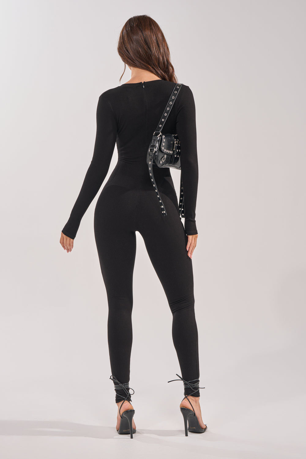 A woman with long brown hair faces away, wearing the DEEP V LONG SLEEVE JUMPSUIT in black ribbed knit, paired with black ankle-strap heels and a small black studded crossbody bag against a plain light gray background. 3