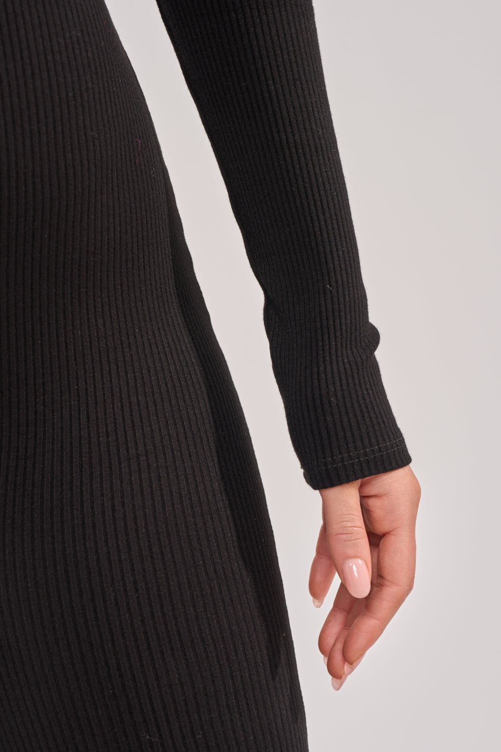 A close-up of a person with light skin and pale manicured nails, resting their arm by their side while wearing the DEEP V LONG SLEEVE JUMPSUIT in black ribbed knit. 5