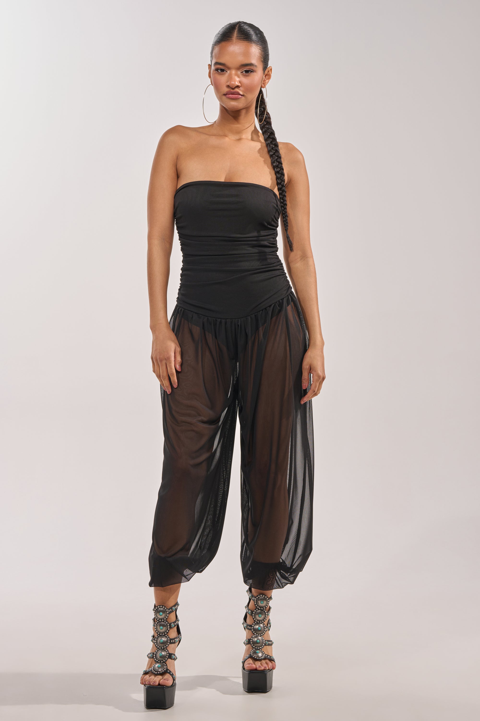 A woman stands confidently in the NO CHILL MESH JUMPSUIT IN BLACK, featuring a strapless mesh bodice and sheer black harem pants with ruched ankles, accessorized with large hoop earrings, braided hair, and platform sandals. 0