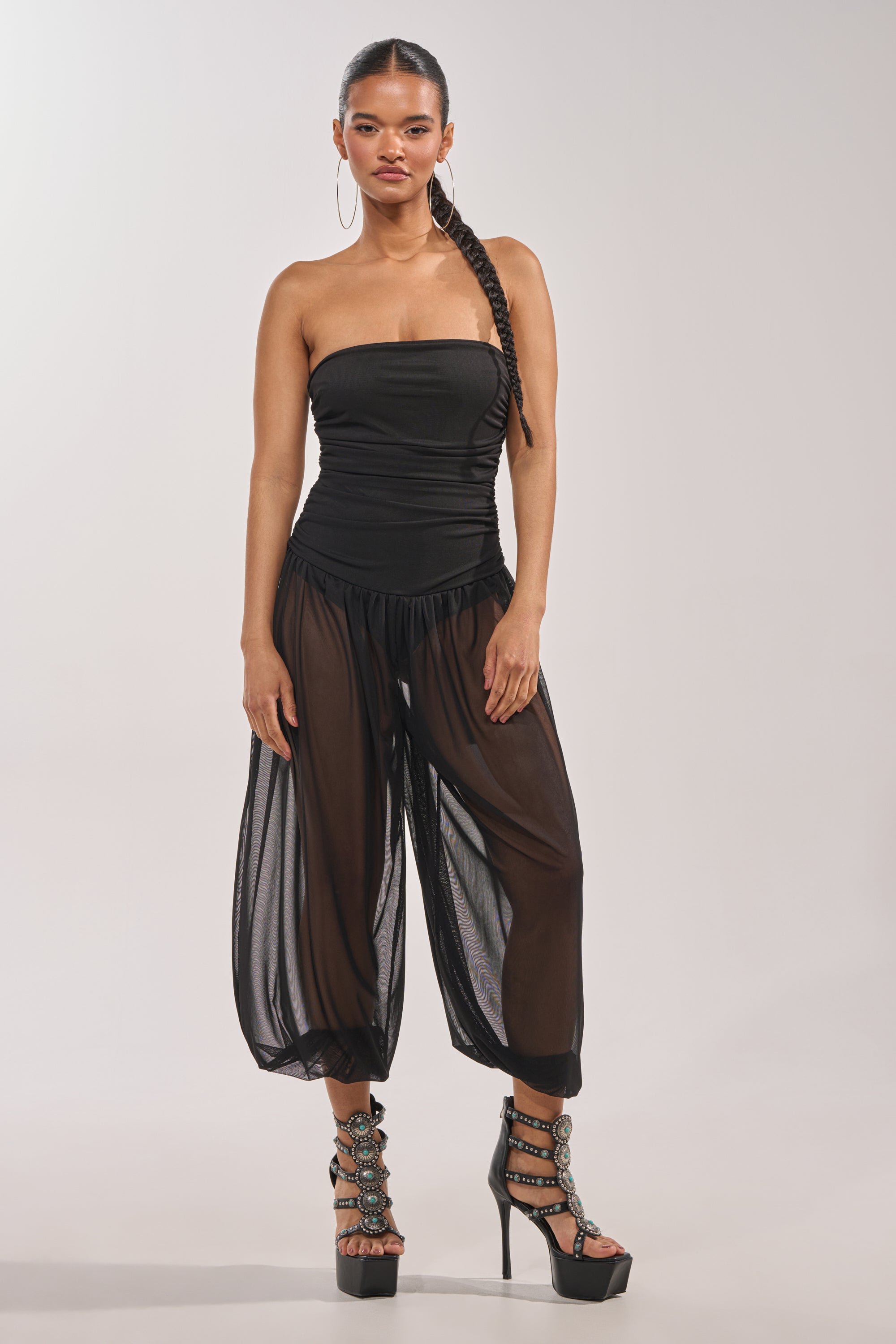 A woman stands confidently against a plain light background, wearing the NO CHILL MESH JUMPSUIT IN BLACK with sheer wide-leg harem pants and high platform heels featuring decorative straps. 1