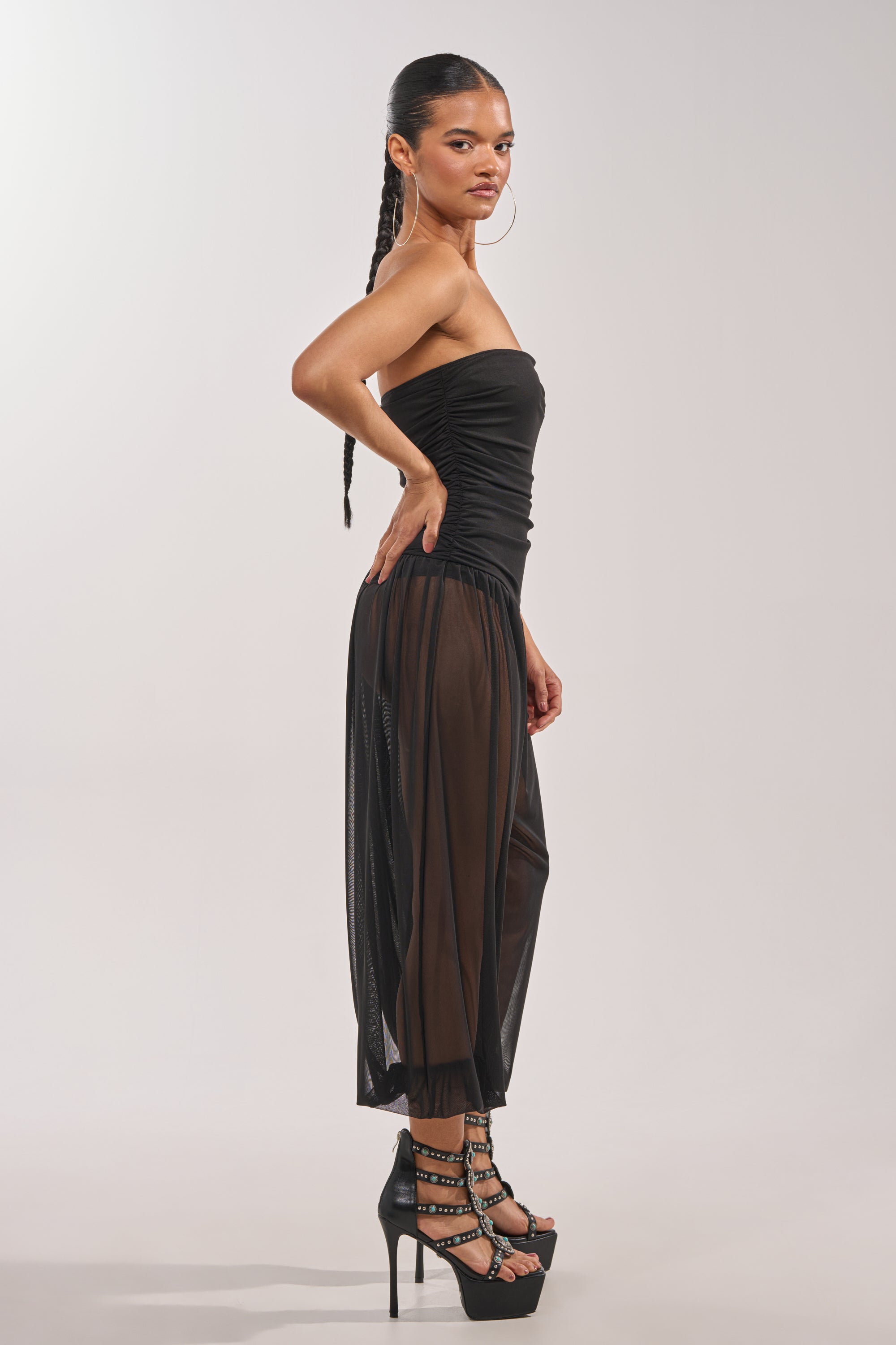 A woman with a long braided ponytail poses sideways against a plain light background, modeling the NO CHILL MESH JUMPSUIT IN BLACK with strappy high-heeled sandals. 2