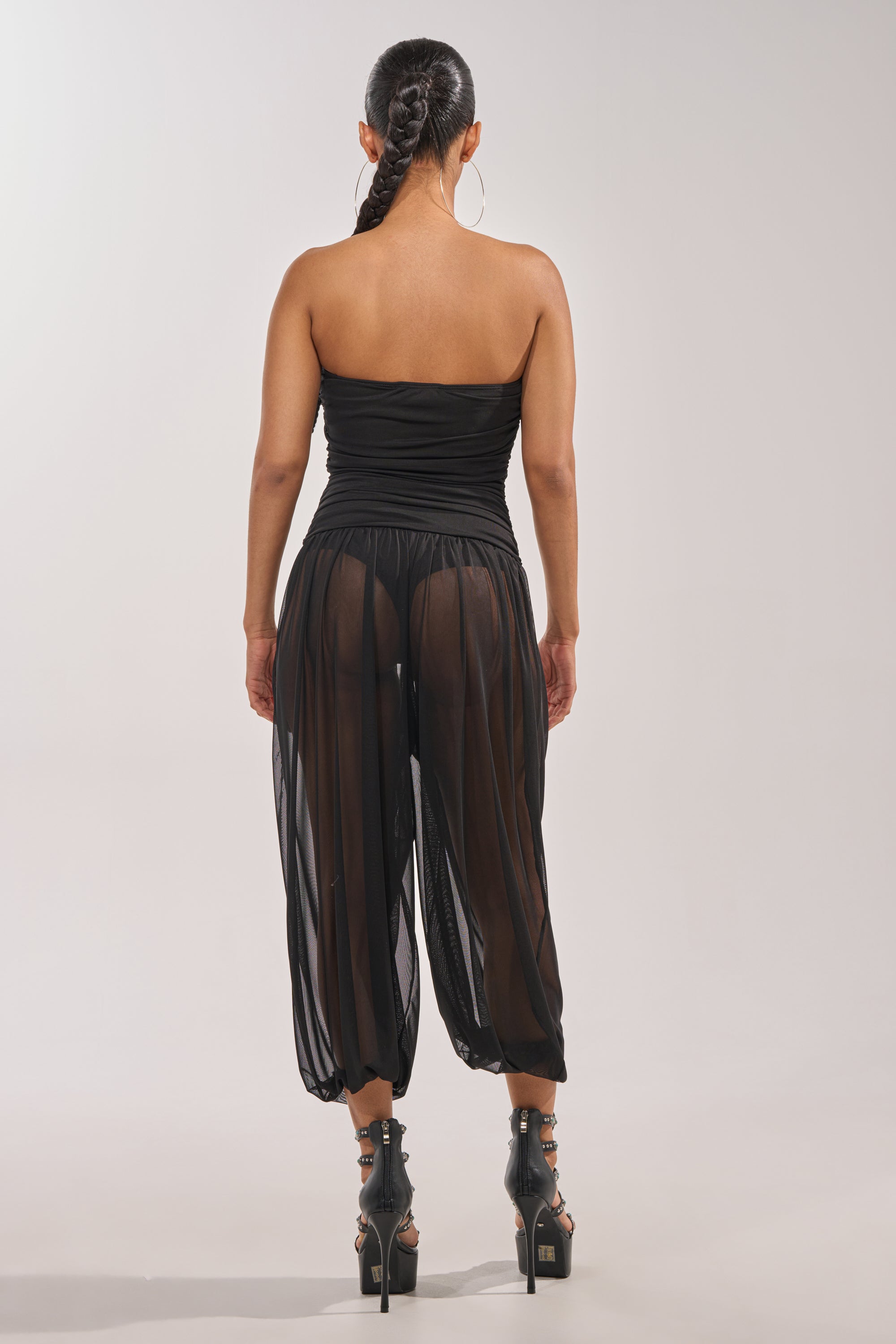 A woman with braided hair faces away, wearing the NO CHILL MESH JUMPSUIT IN BLACK with visible black shorts and high-heeled sandals—an ideal evening jumpsuit against a plain background. 3