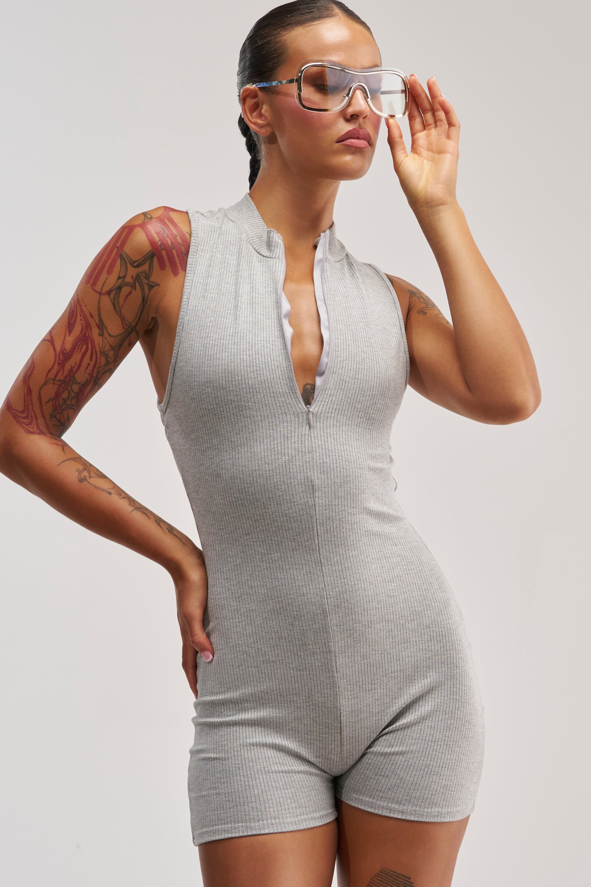 A woman models the EFFORTLESS ICON ZIP UP KNIT ROMPER—a sleeveless, grey rib knit piece—paired with clear visor glasses. With one hand on her hip and the other adjusting her glasses, her tattooed arms and confident pose bring a bold streetwear vibe to the neutral backdrop.