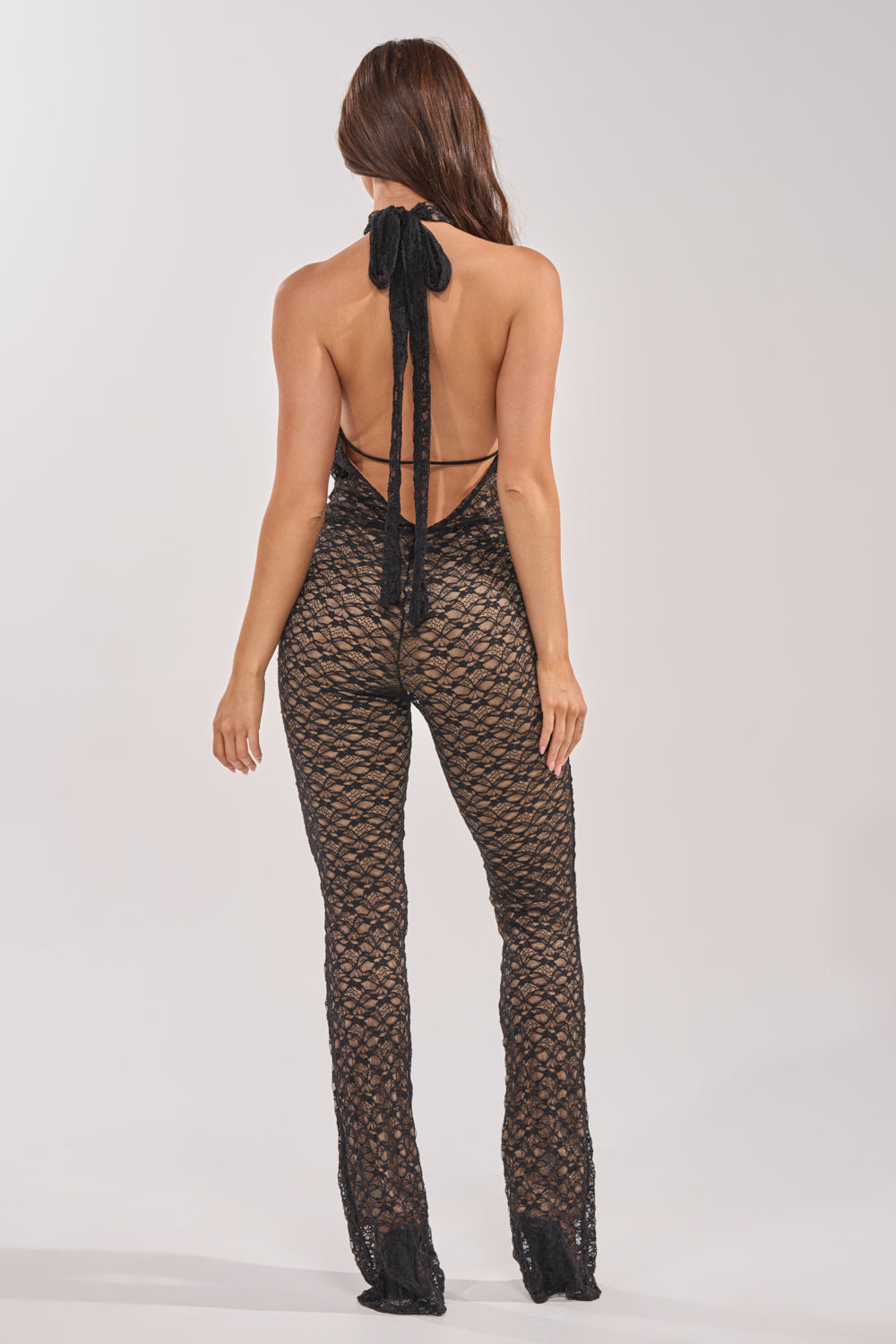 A woman faces away, wearing the MAIN VIBE LACE JUMPSUIT IN BLACK featuring a halter neck and flared legs—an effortlessly chic choice for a girls’ night out. Her long brown hair falls loose against a plain, light background. 4