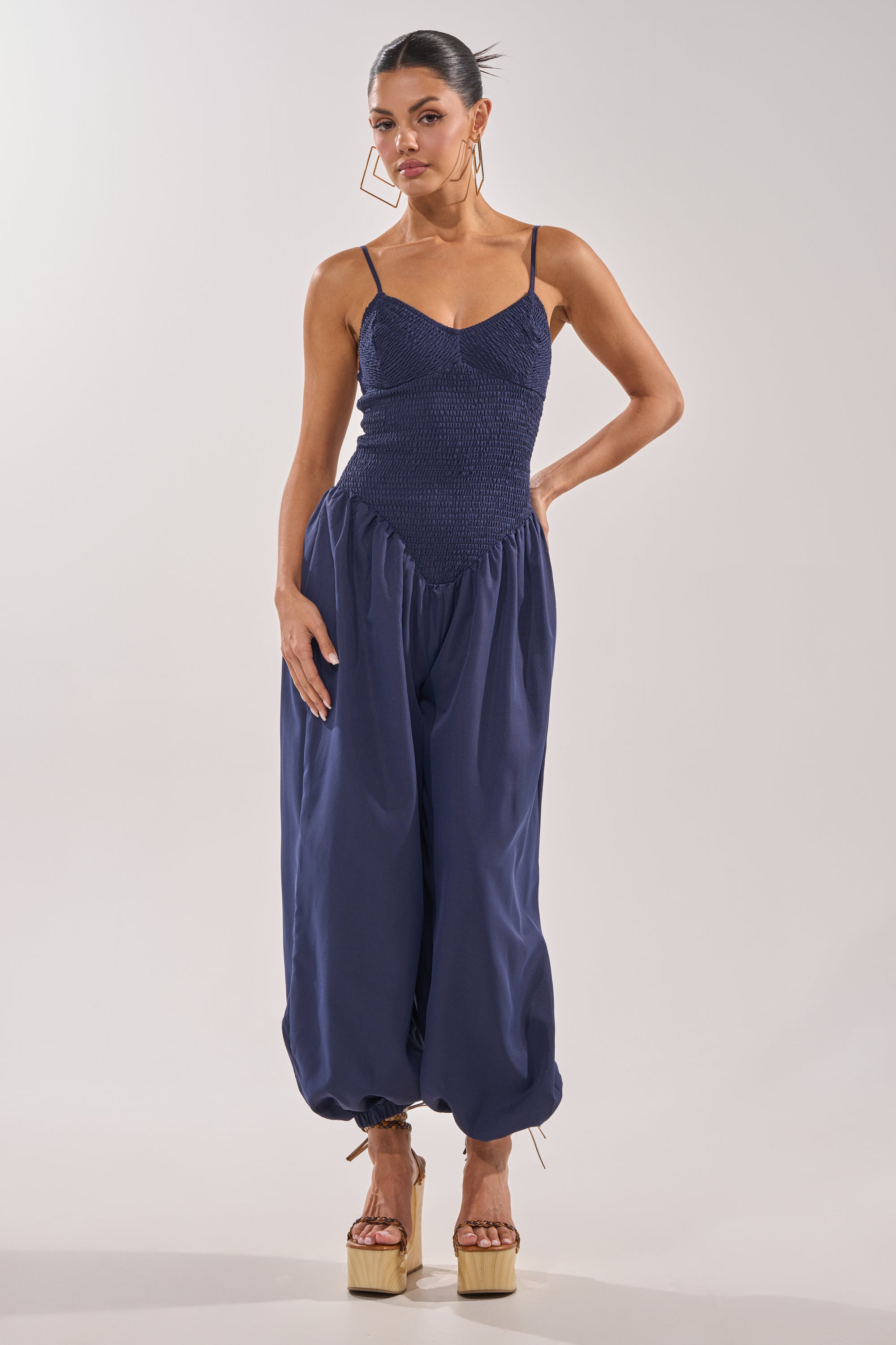 A woman wears the TRENDSETTER BALLOON PANT JUMPSUIT, featuring a smocked navy bodice, spaghetti straps, and loose pants gathered at the ankles. She completes her chic look with geometric earrings and platform sandals. 1
