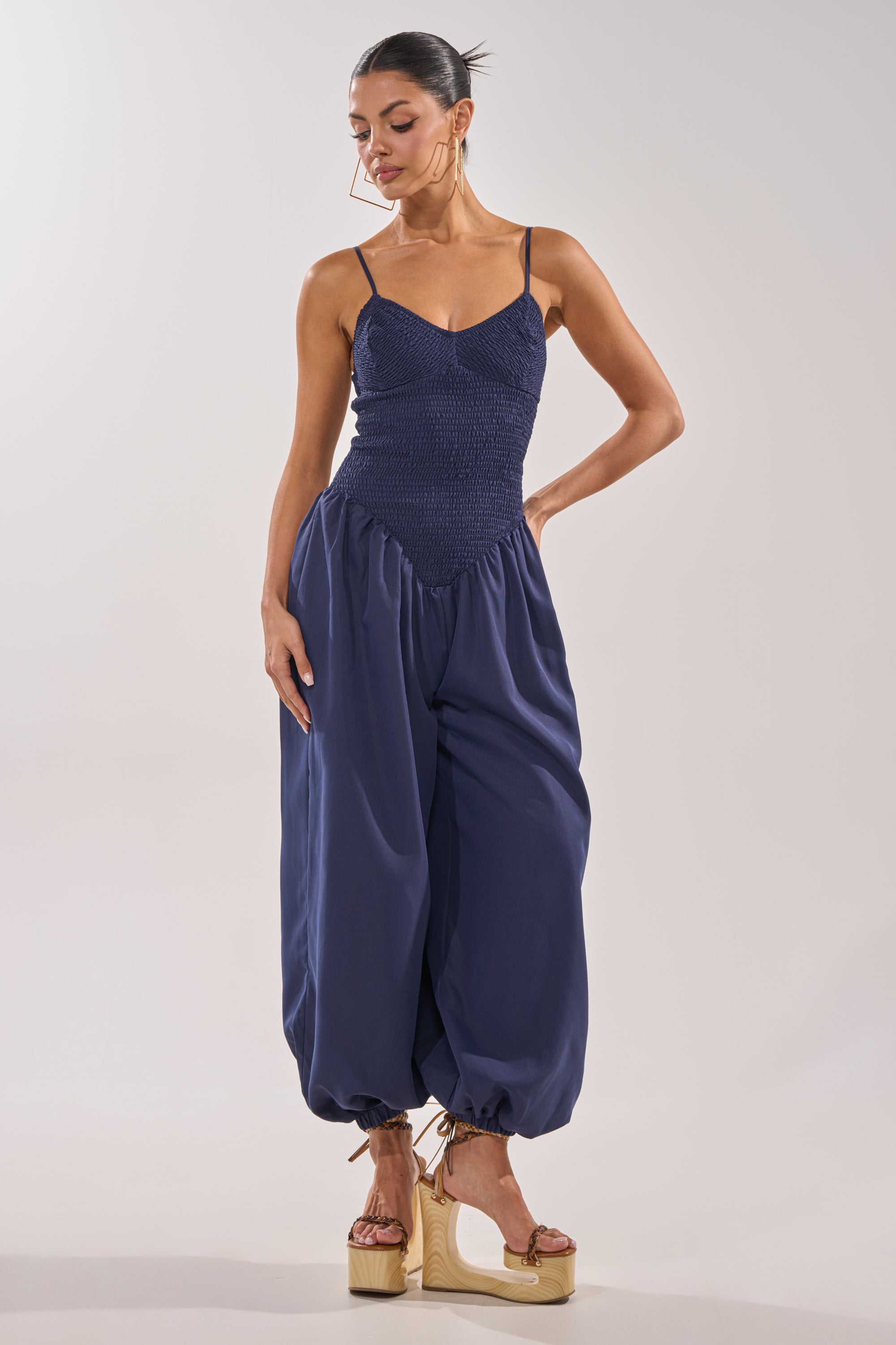 A woman wears the TRENDSETTER BALLOON PANT JUMPSUIT—a navy smocked style with a ruched bodice and billowy, ankle-gathered pants—paired with tan platform sandals, standing against a plain light background looking down and to the side. 2