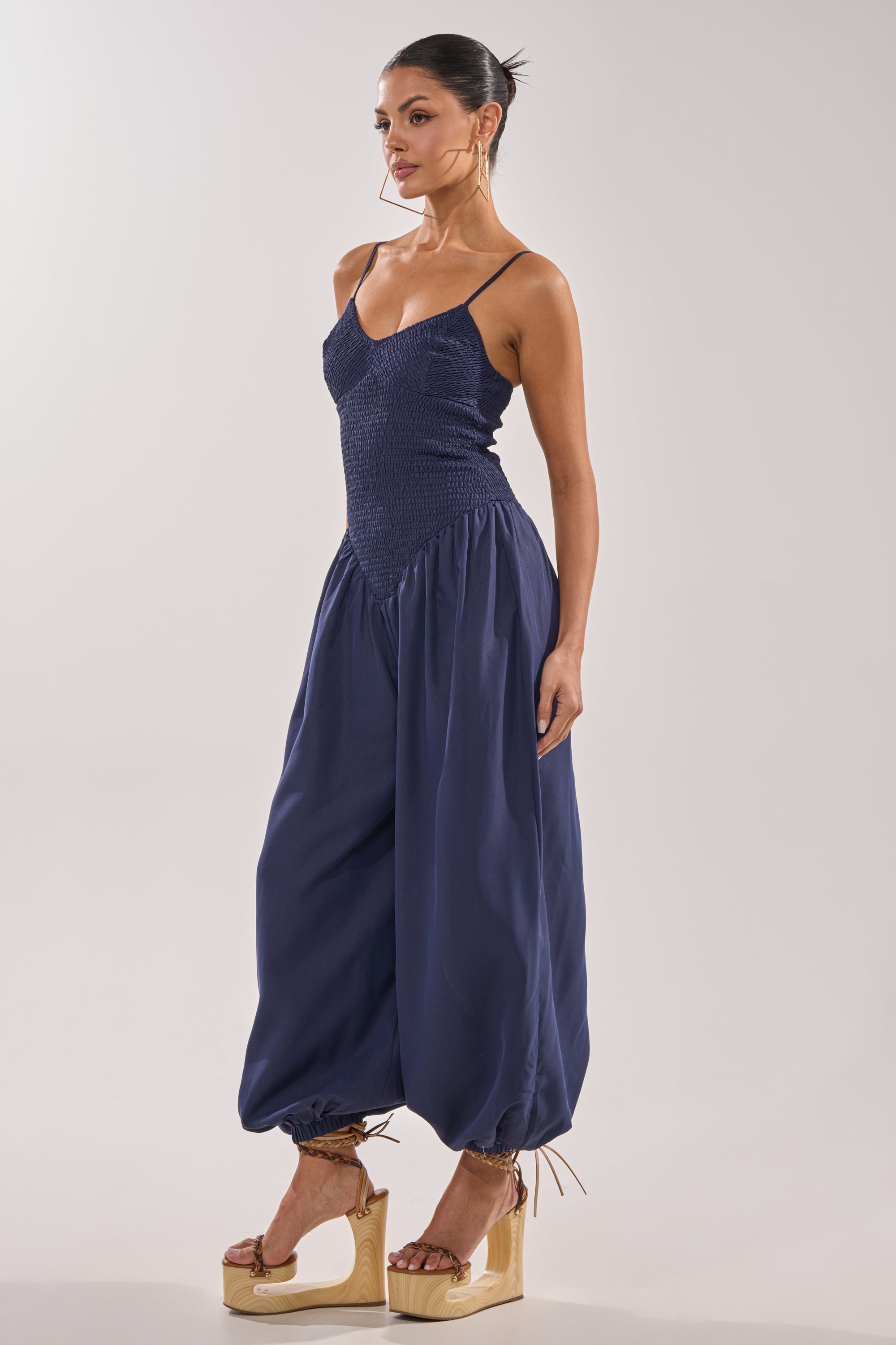 A woman in profile wears the TRENDSETTER BALLOON PANT JUMPSUIT, featuring a smocked navy bodice and loose, gathered legs cinched at the ankles. Her hair is up, styled with tan platform sandals for a chic evening look. 3