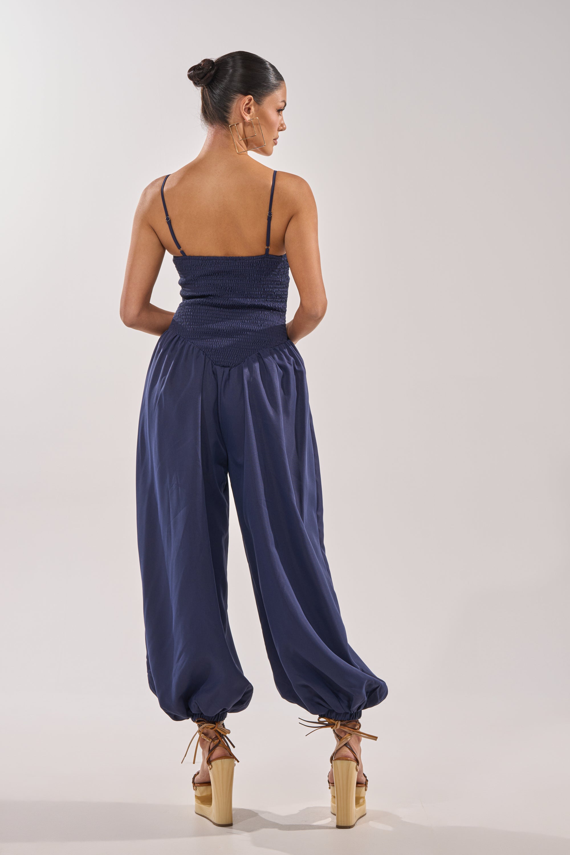 A woman with her hair in a bun faces away, wearing the TRENDSETTER BALLOON PANT JUMPSUIT—navy, smocked bodice, spaghetti straps, loose balloon pants cinched at the ankles—paired with tan platform heels against a plain light background. 4
