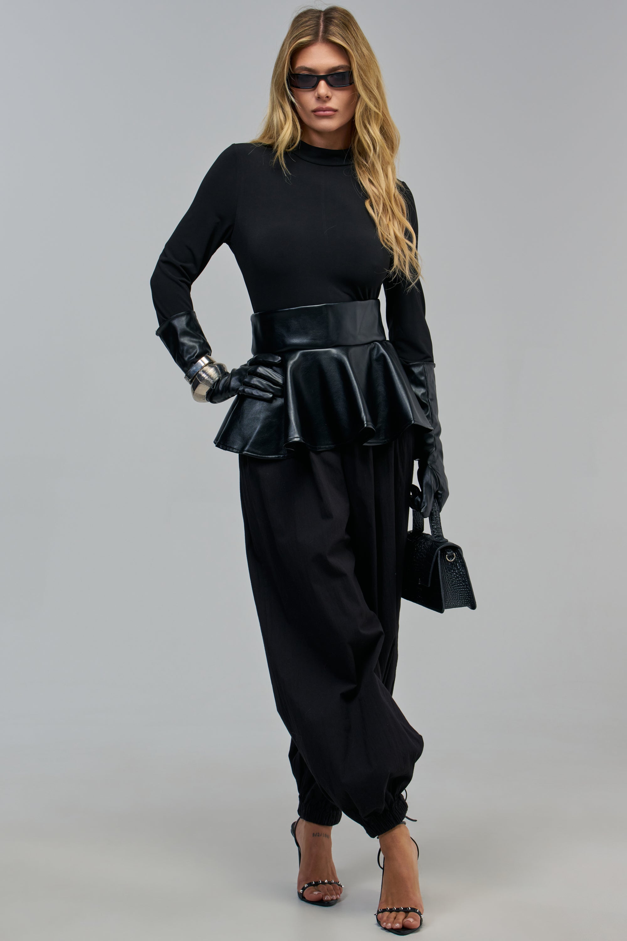 A model wears the PROFESSIONAL BADDIE PEPLUM JUMPSUIT IN BLACK, a long-sleeved black peplum jumpsuit, styled with leather gloves and strappy sandals for an effortless workwear look.