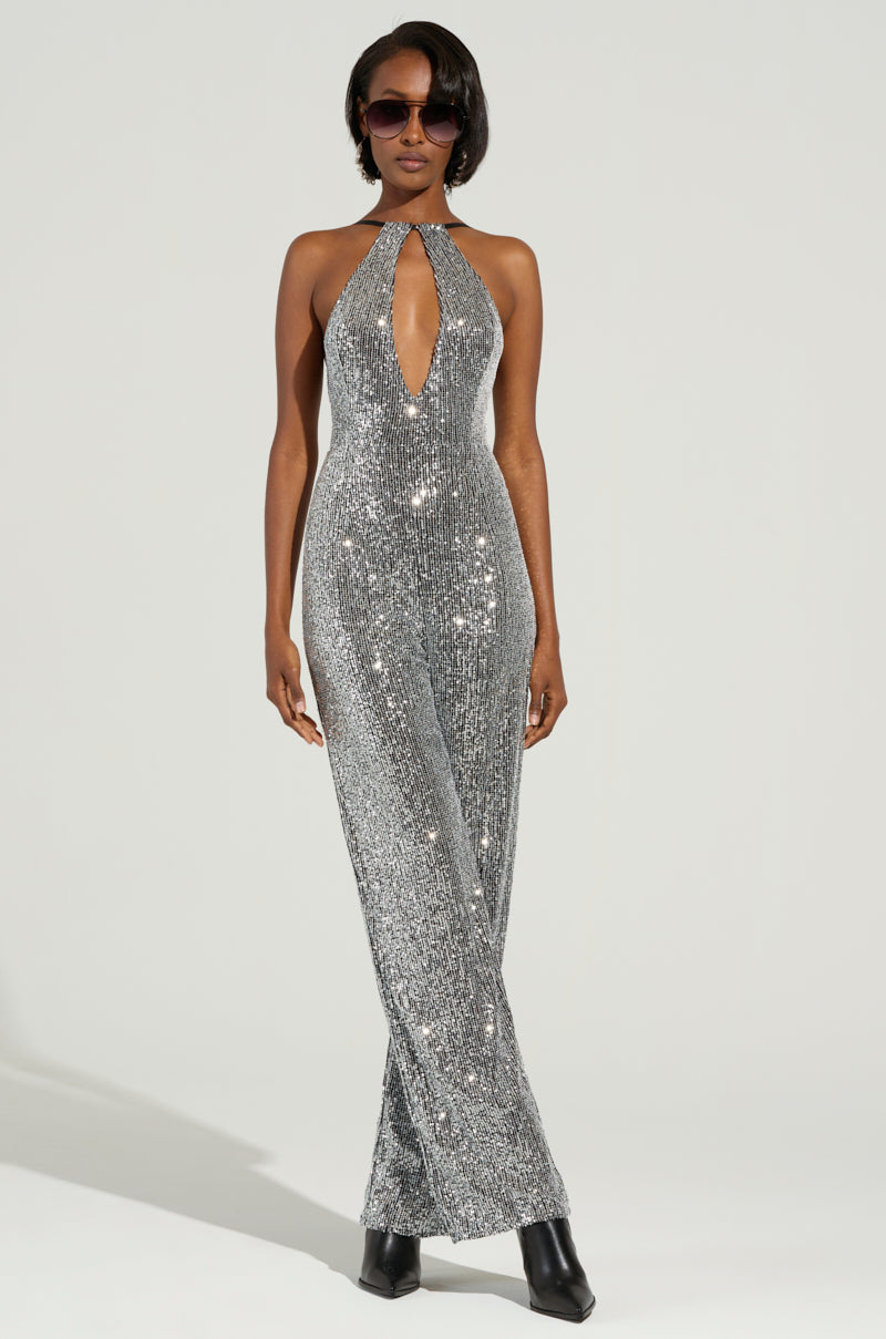A person is wearing the CAN'T MISS ME SEQUIN JUMPSUIT, sparkling in silver sequins with a deep halter neckline, paired with black heeled boots. They have short hair and are accessorized with dark sunglasses while posing against a plain backdrop.