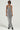 A person is wearing the CAN'T MISS ME SEQUIN JUMPSUIT, sparkling in silver sequins with a deep halter neckline, paired with black heeled boots. They have short hair and are accessorized with dark sunglasses while posing against a plain backdrop.