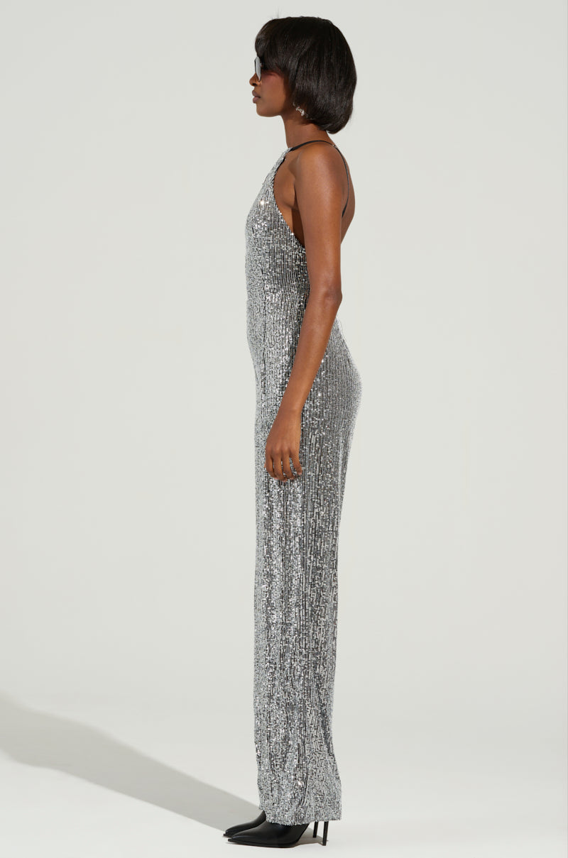 A woman in the eye-catching "CAN'T MISS ME SEQUIN JUMPSUIT" poses in profile against a plain backdrop. Her short dark hair is chicly styled, and she accents her look with sunglasses and sleek black heeled shoes for an elegant finish.