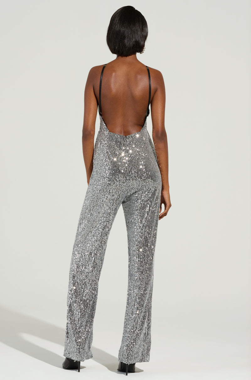 A person stands facing away, wearing the CAN'T MISS ME SEQUIN JUMPSUIT that features a halter neckline and low back. The sparkling silver sequins and rhinestone embellishments catch the light as they pose confidently, with short dark hair contrasting against the plain light backdrop.