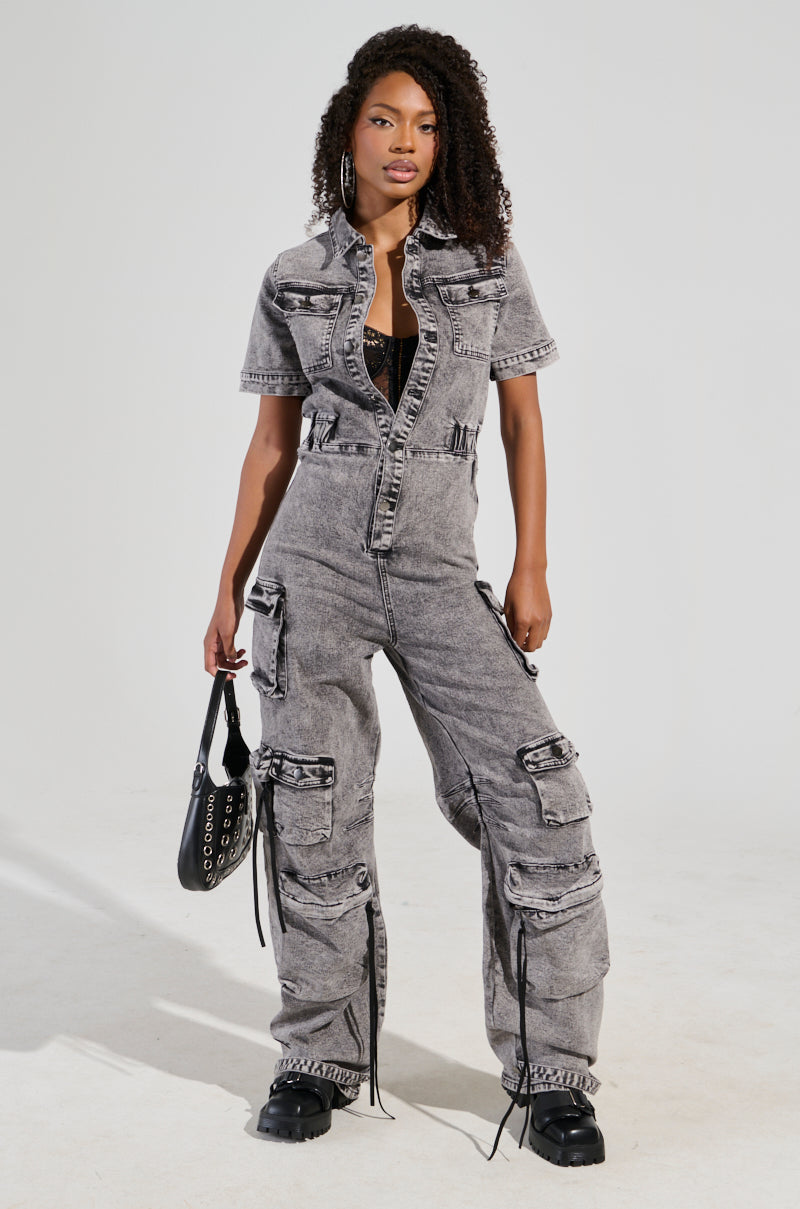 Wearing the UTILITY CARGO DENIM JUMPSUIT IN GREY, a person stands confidently with its wide-leg silhouette and multiple pockets. They pair it with a black top and boots, accessorized with hoop earrings and a black handbag, as their curly hair cascades over one shoulder.