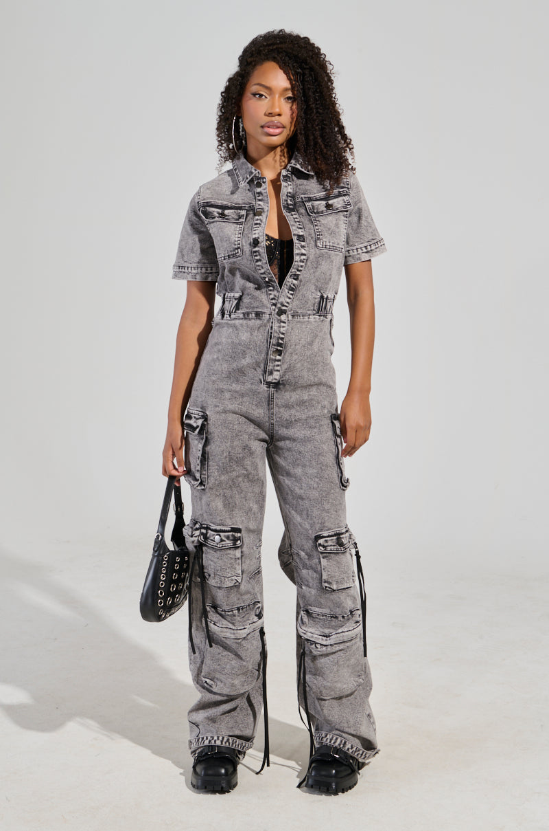 A woman with curly hair is wearing the utility cargo denim jumpsuit in grey, which features a wide leg silhouette and large pockets, paired with black boots. She holds a black handbag adorned with circular decorations, standing against a plain background.