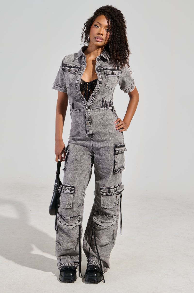 A person stands confidently wearing the Utility Cargo Denim Jumpsuit in Grey, featuring multiple pockets, over a patterned black top. They have long, curly hair and are holding a black handbag, set against a simple and light background.