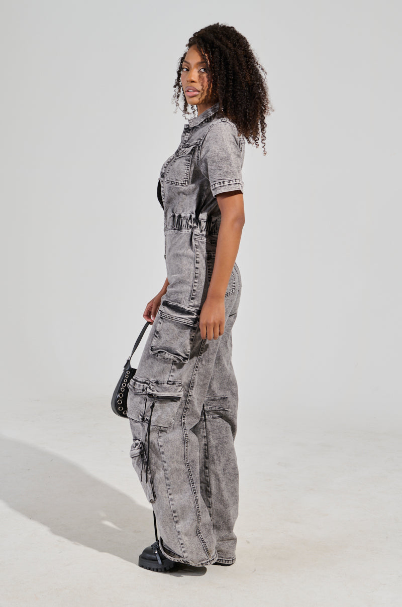 A person with curly hair is seen standing sideways in a Utility Cargo Denim Jumpsuit in grey, showcasing pockets and zippers that highlight its utility style. They are holding a small black bag and wearing black boots, all set against a plain, light-colored background.