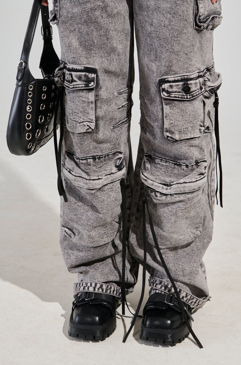Close-up of a person wearing the Utility Cargo Denim Jumpsuit in grey, featuring a wide-leg silhouette, distressed details, multiple pockets, and black buckled shoes. They are holding a black handbag with silver grommets, while black straps hang from the jumpsuit.