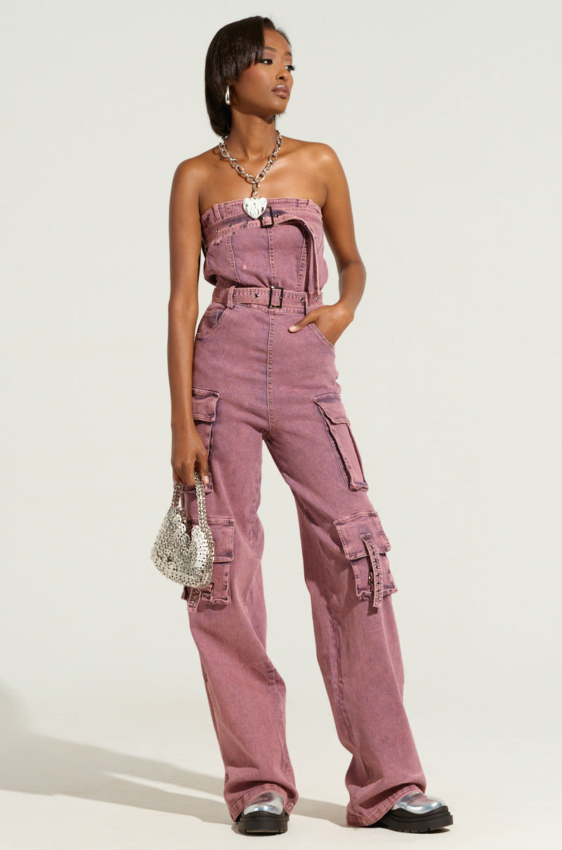 A person stands confidently in a stylish acid washed denim jumpsuit with a wide-leg silhouette and multiple pockets, wearing a silver chain necklace. They hold a small textured handbag and have shiny black shoes. The background is a neutral, soft tone.