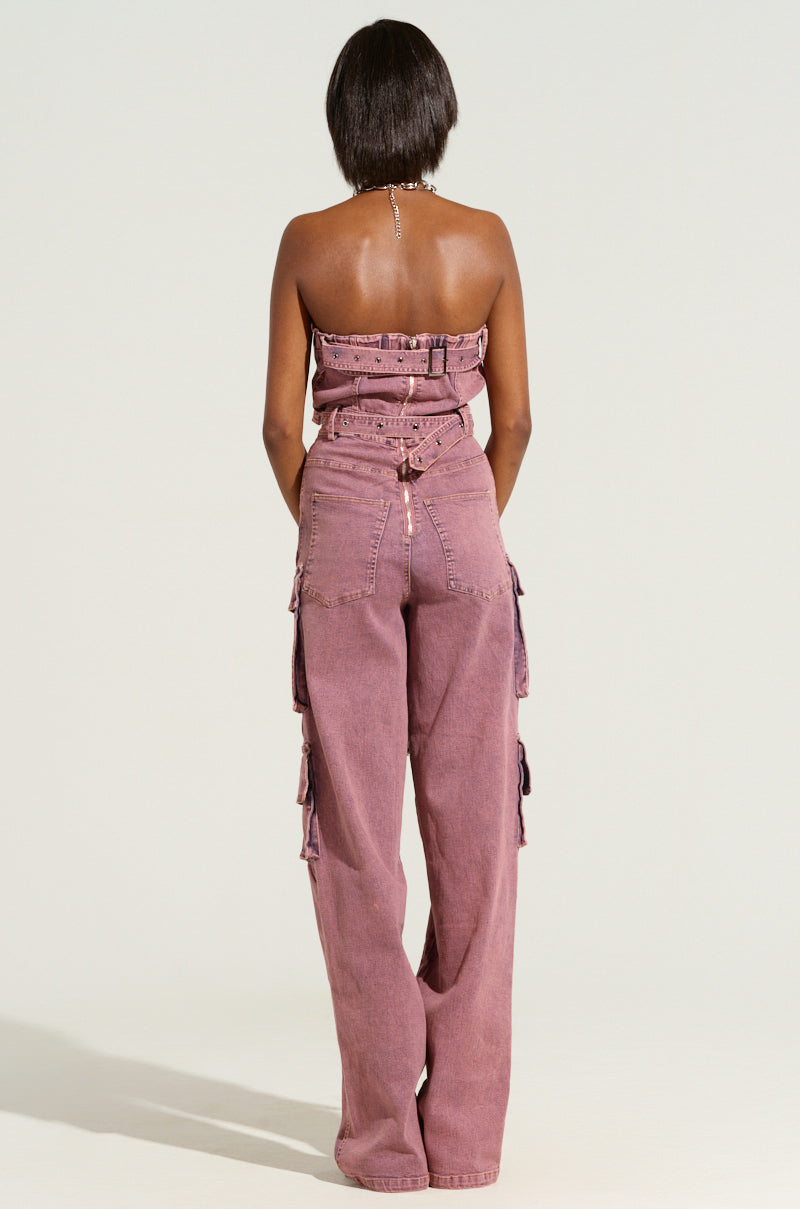 A person with short dark hair is facing away from the camera, wearing an ACID WASHED DENIM JUMPSUIT in a faded purple color. The jumpsuit includes several cargo pockets and a belt around the waist.