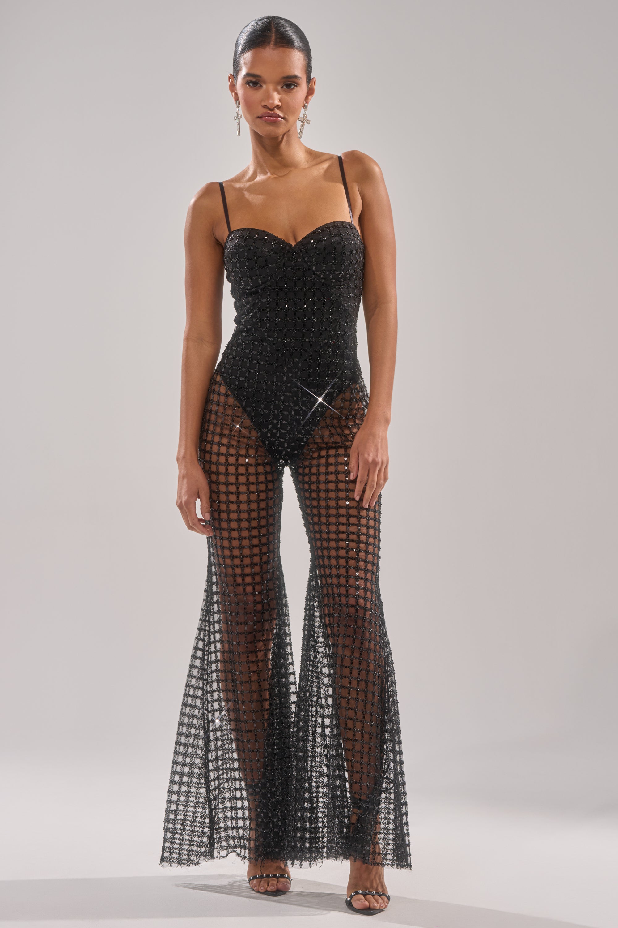 A woman models the HARDEST TO LOVE MESH JUMPSUIT—black, strapless, with sheer flared pants, a checkered pattern, and sparkling sequin accents—set against a plain light background. 0