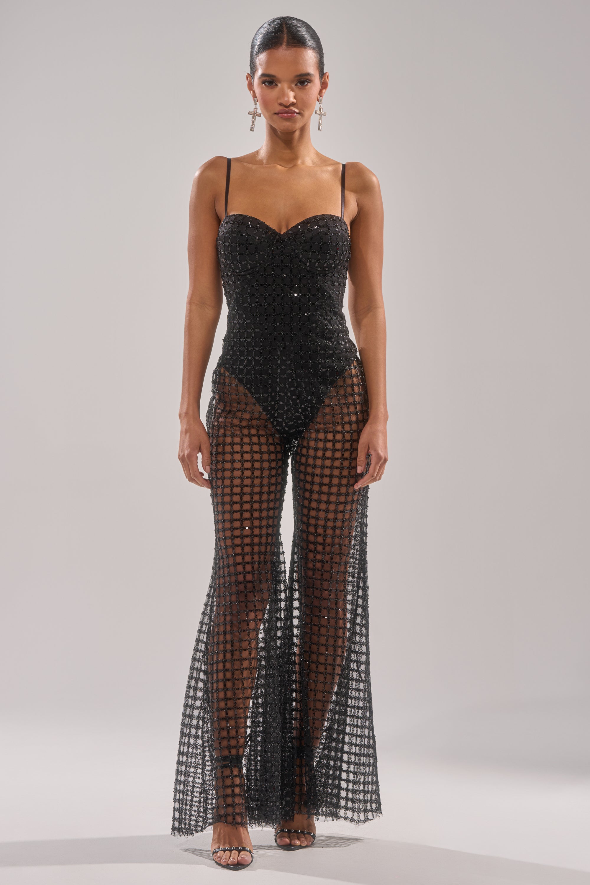 A woman faces forward in the HARDEST TO LOVE MESH JUMPSUIT—a black strapless piece with a sheer, floor-length checkered overlay. With slicked-back hair, dangling earrings, and open-toe heels, it’s perfect for a chic night out. 2