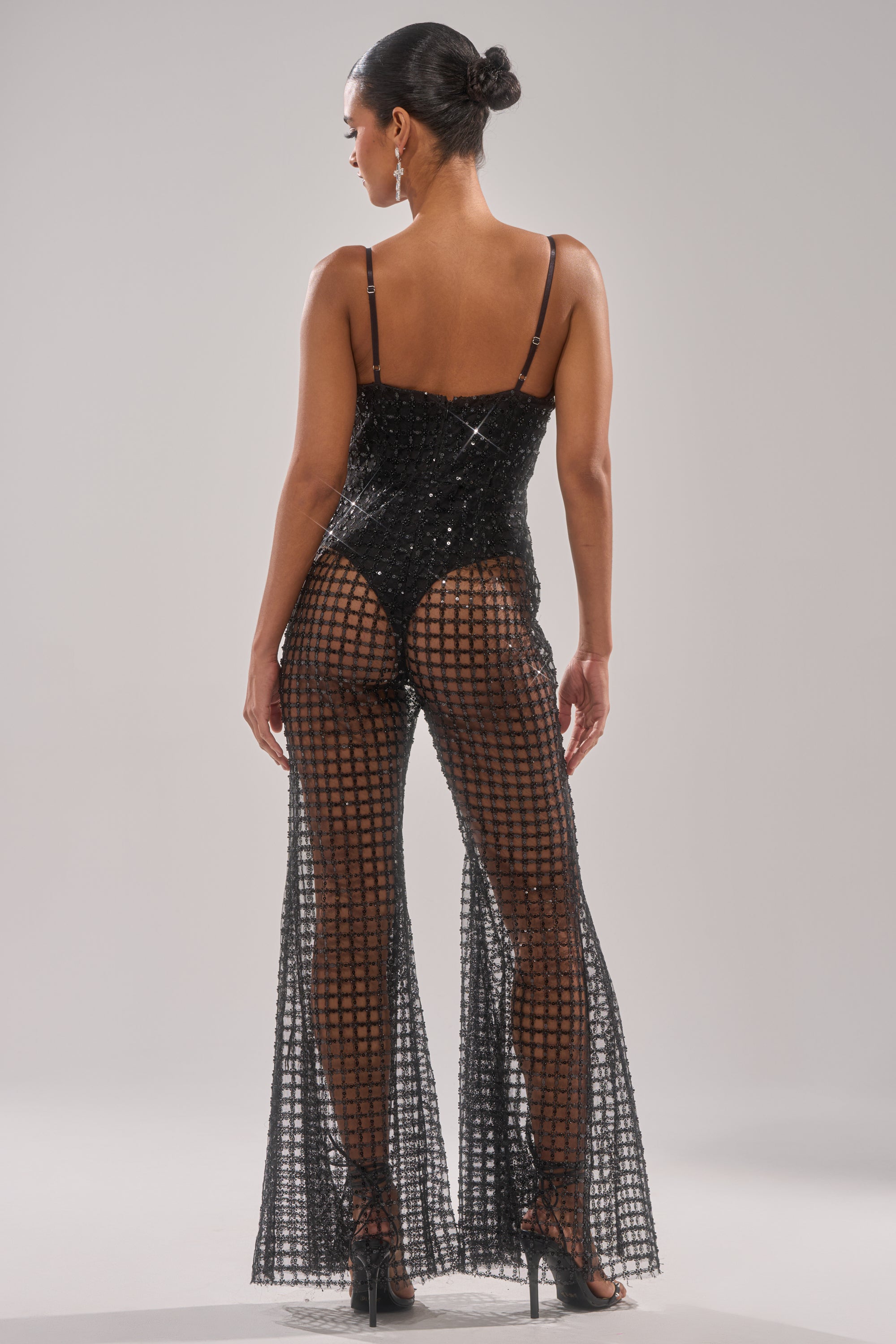 A woman faces away, wearing the HARDEST TO LOVE MESH JUMPSUIT—sparkly black mesh with flared legs—paired with strappy heels. Her hair is in a sleek bun, accessorized by dangling earrings. 4