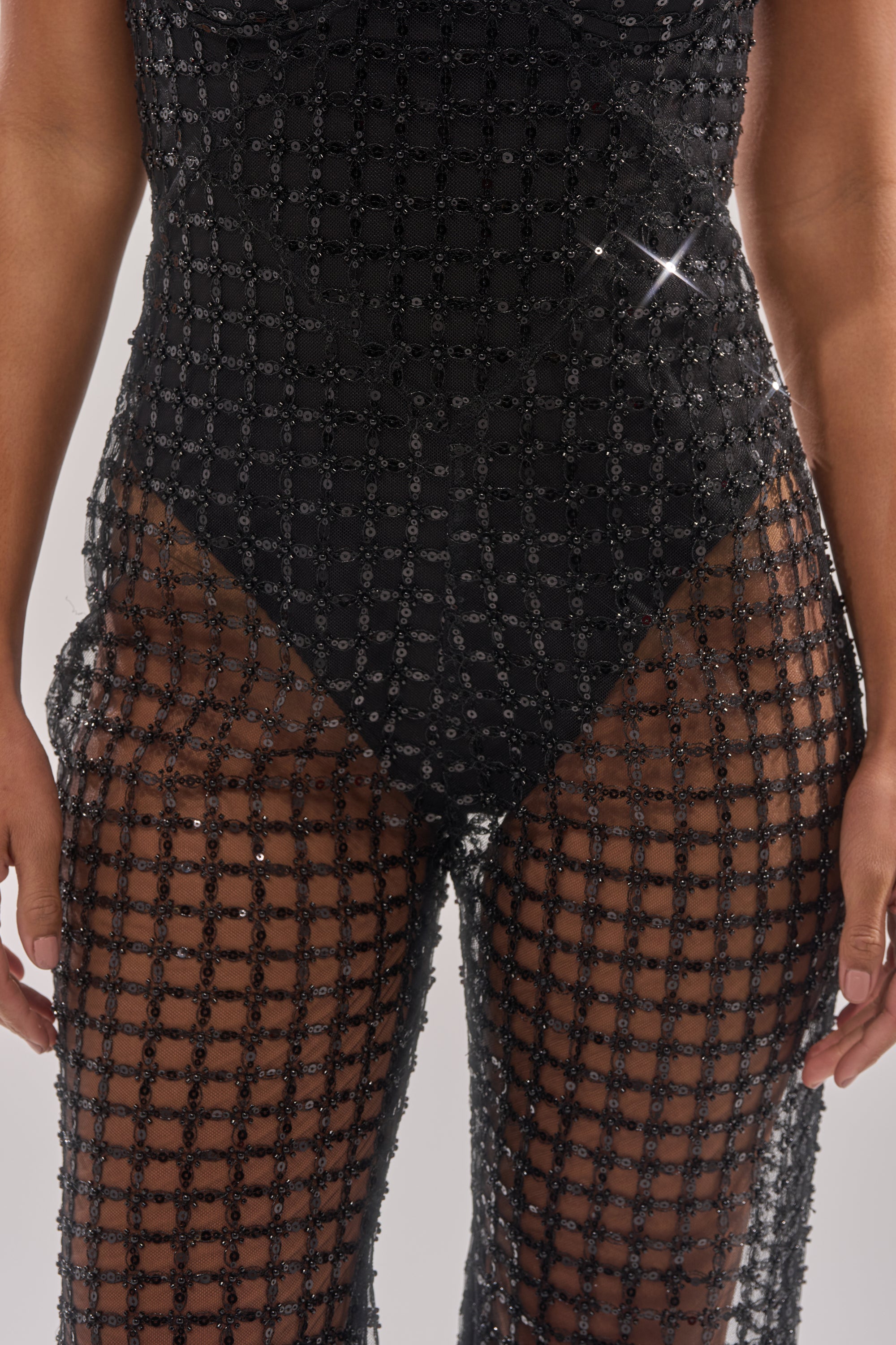 A close-up of a person wearing the HARDEST TO LOVE MESH JUMPSUIT, featuring black mesh with a geometric grid pattern and sequin accents. The sheer design reveals skin on the arms and midsection. 5