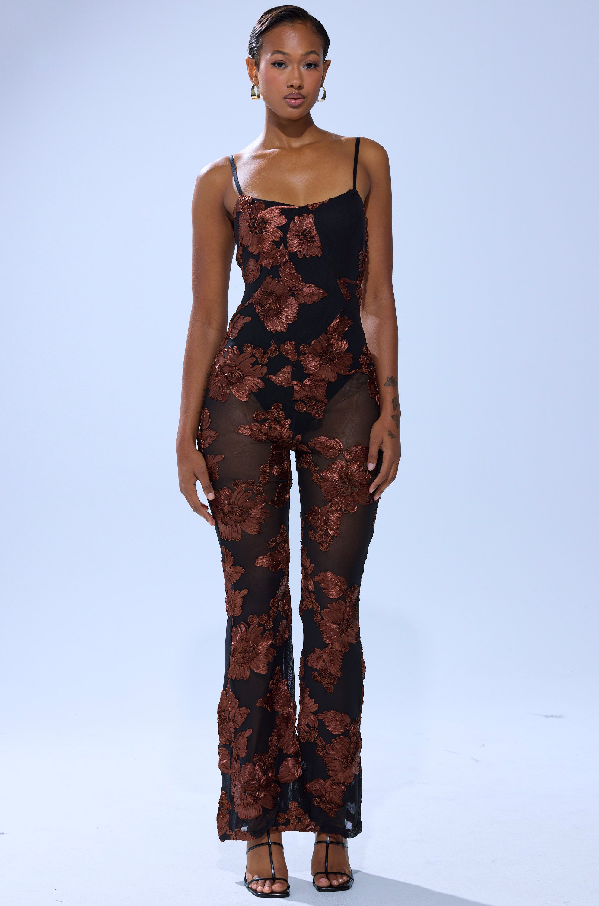 A woman faces forward in the LAY IT LOW PRINTED MESH JUMPSUIT, featuring large brown floral prints, sheer black mesh, thin straps, and black high-heeled sandals against a plain light background.