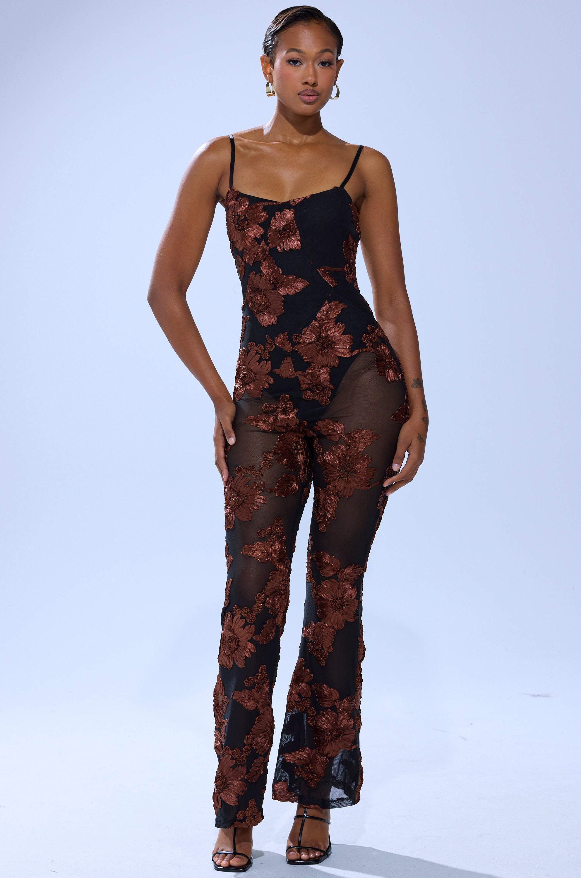 A woman stands confidently in the LAY IT LOW PRINTED MESH JUMPSUIT, featuring brown floral patterns and thin straps, paired with black heels against a plain light background.
