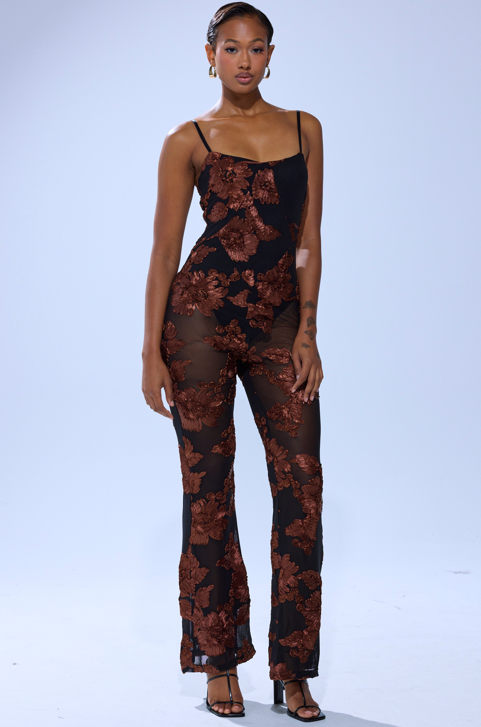 A woman poses facing forward in the LAY IT LOW PRINTED MESH JUMPSUIT, a sheer black jumpsuit with bold brown floral prints, paired with black heels and hoop earrings against a plain background.