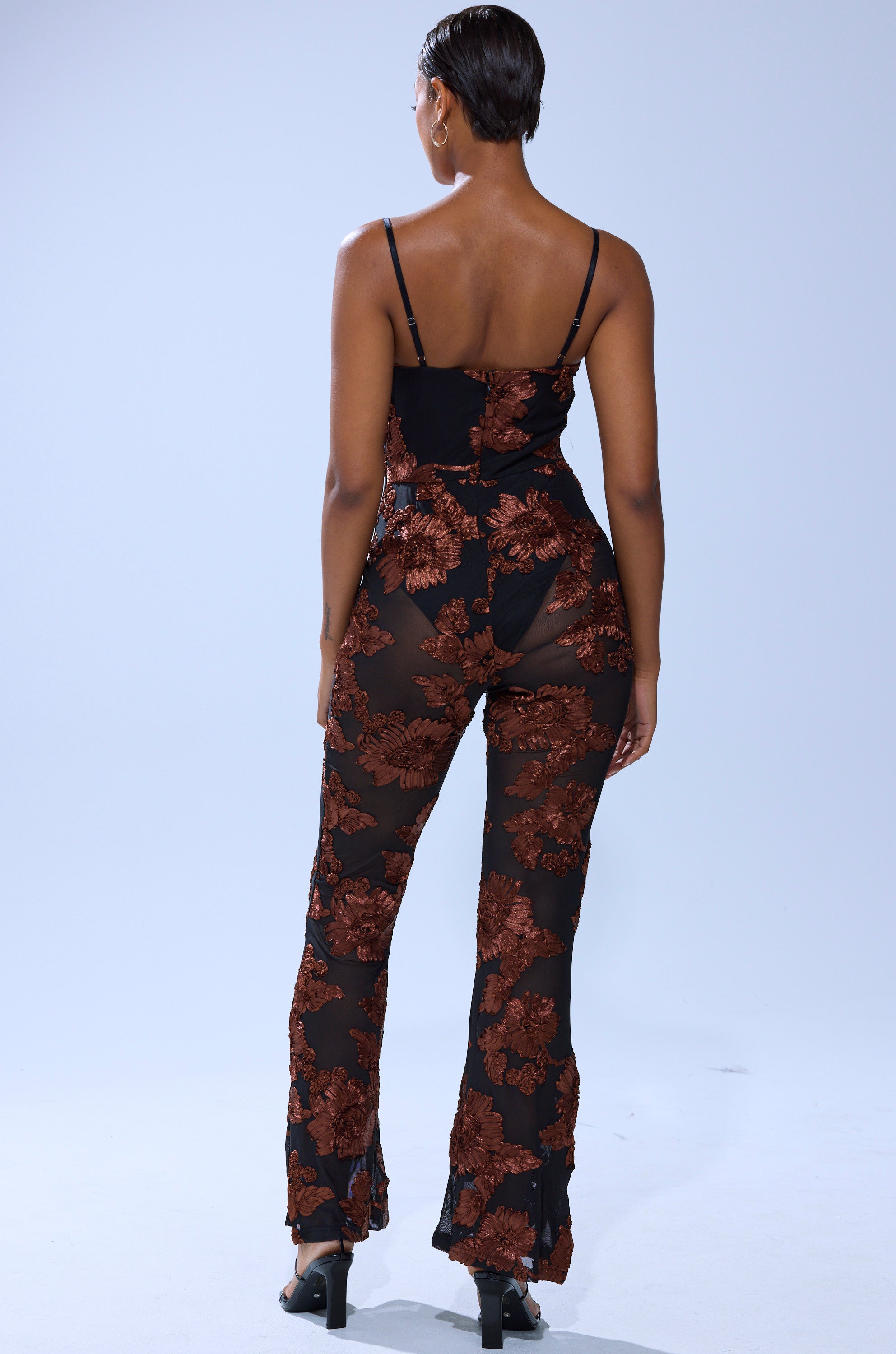 A person stands facing away, wearing the LAY IT LOW PRINTED MESH JUMPSUIT with brown floral patterns and black heels, against a plain light background.