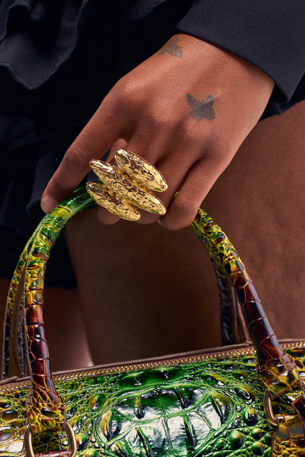 A close-up shot shows a hand with butterfly tattoos on the fingers holding the green and brown crocodile-embossed handle of a handbag. The hand wears the STACK IT RING, an ornate, hammered gold stretch ring featuring three prominent, elongated oval shapes.