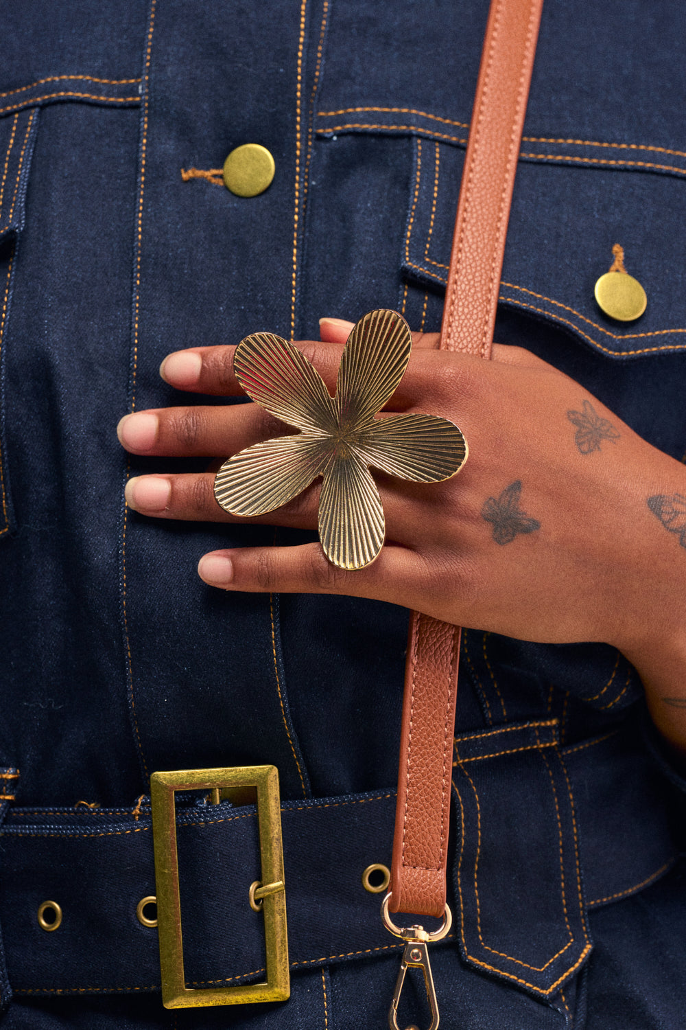 A hand adorned with butterfly tattoos wears the MAKING A STATEMENT RING, a striking gold engraved flower band, while holding a brown strap. The person is dressed in a dark denim outfit accented by gold buttons and a large gold belt buckle.