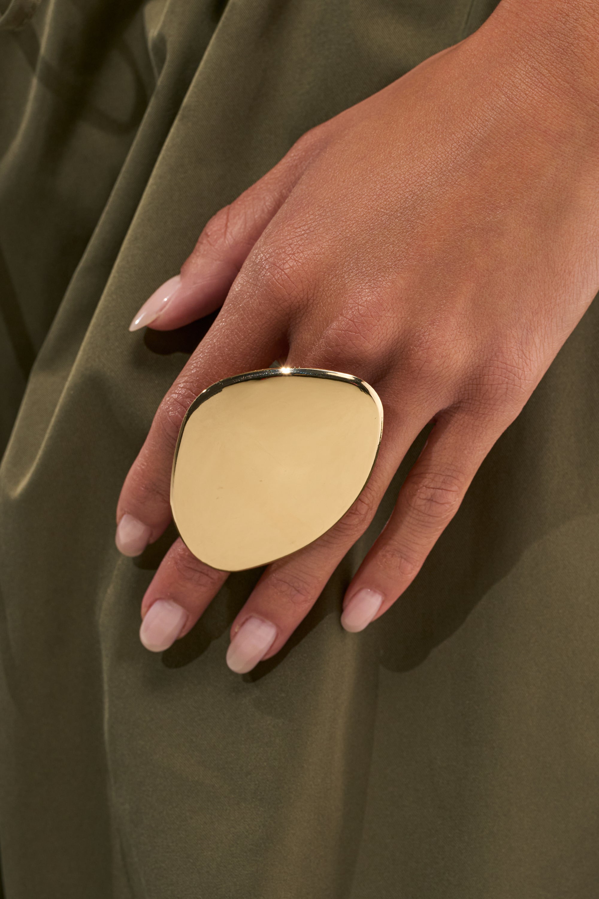 A hand with manicured nails wears the MOD GAL STATEMENT RING—a large, sculptural piece in metallic gold—resting against olive green fabric.