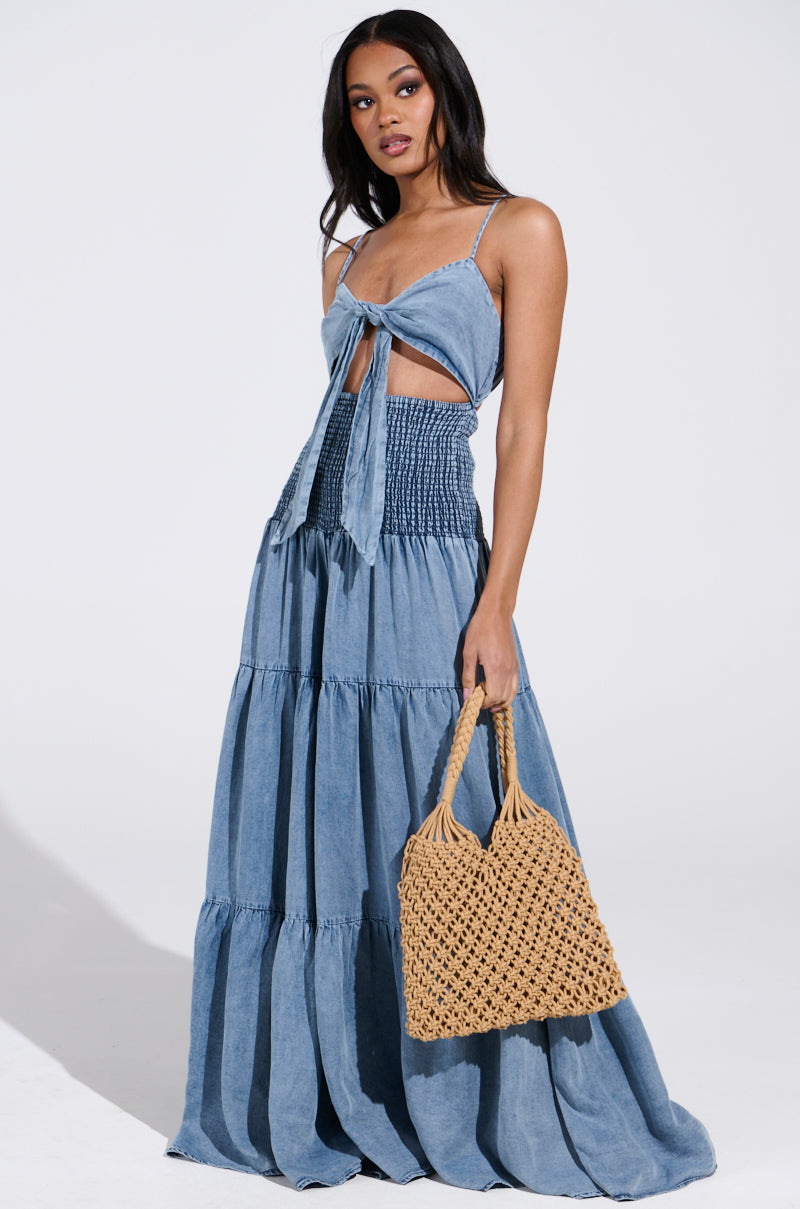 A woman in a JUMPIN AROUND JUMPSUIT, featuring a knotted top and tiered skirt detail, holds a brown woven handbag. She stands against a plain background, looking towards the camera.