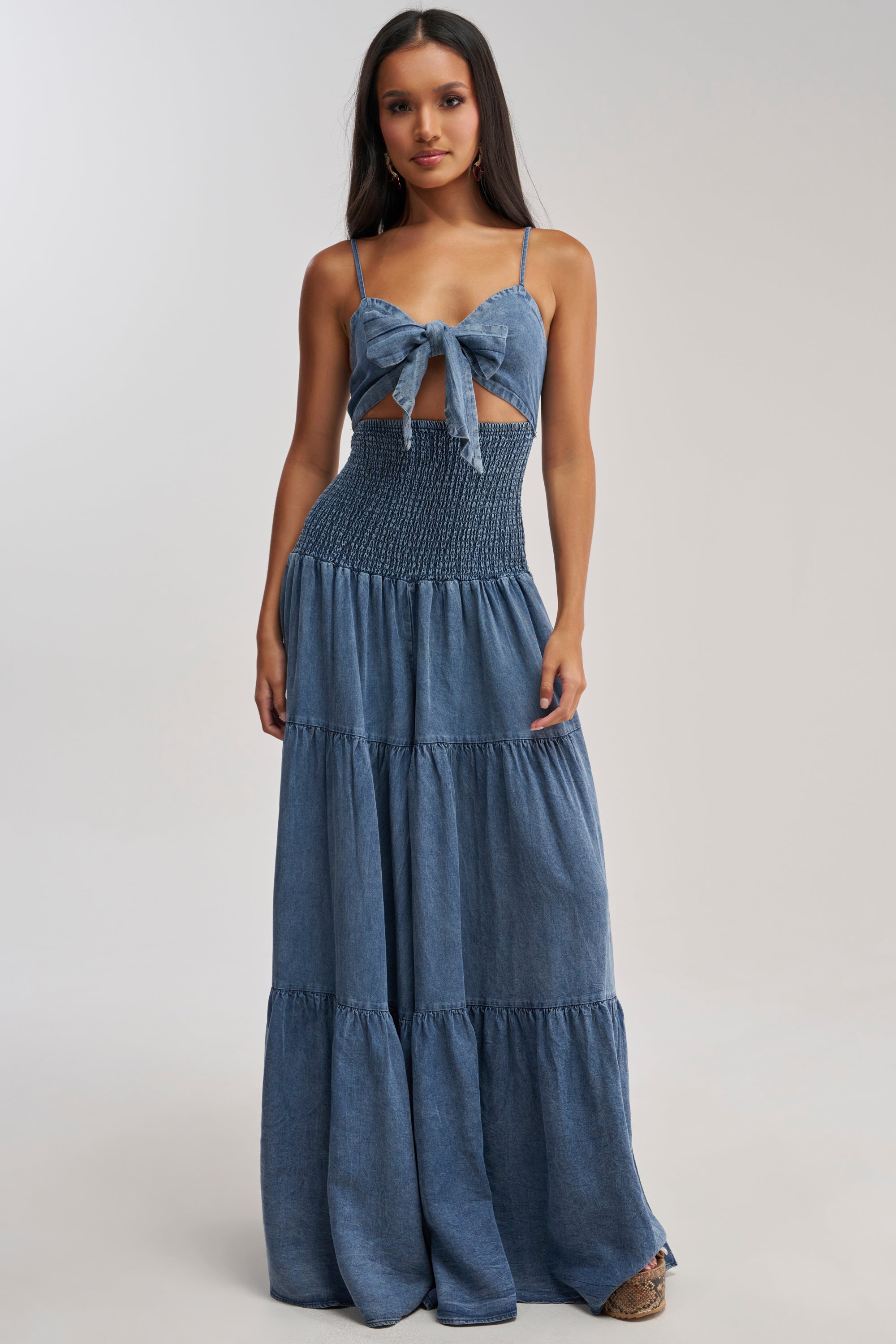 A woman models the JUMPIN AROUND DENIM JUMPSUIT, a sleeveless denim outfit with thin straps, front tie detail, smocked waist, and tiered legs—perfect for vacation—against a plain light background.