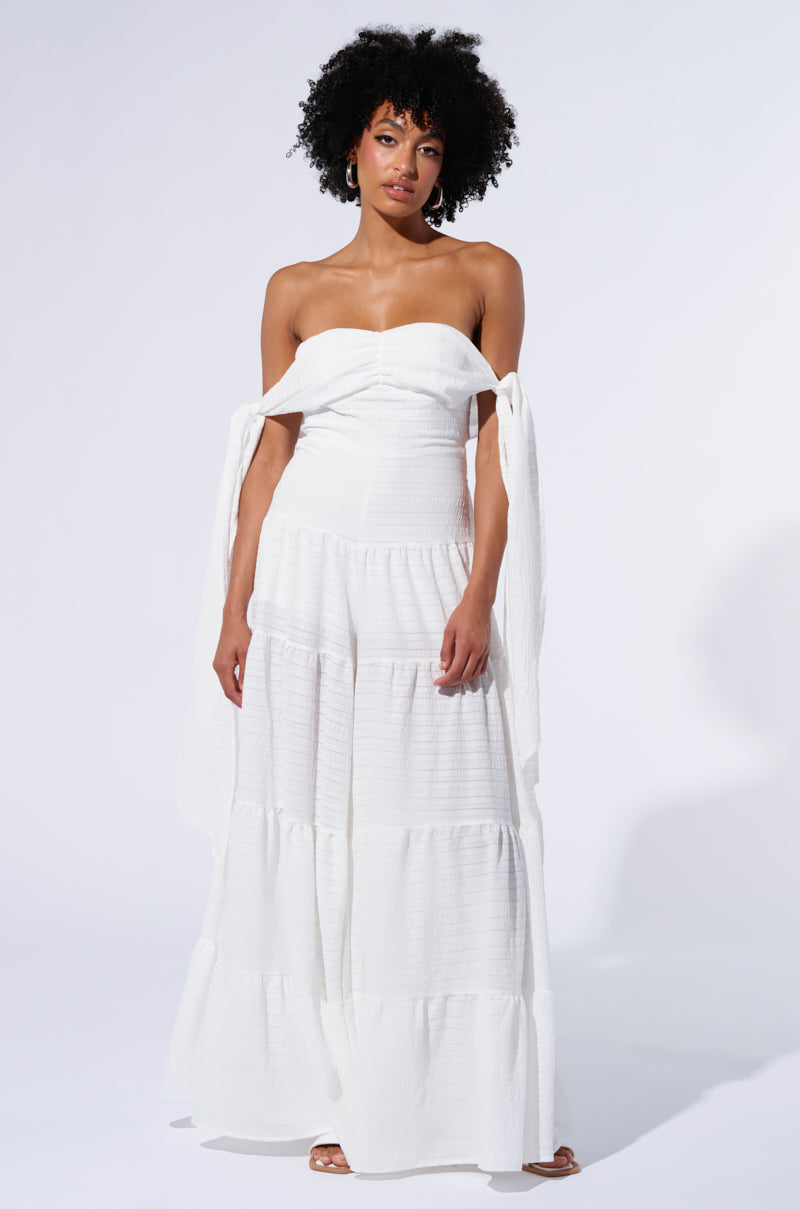 A woman stands against a plain background wearing the GO OFF JUMPSUIT, featuring tiered layers and seersucker detailing. The jumpsuit has tied sleeves hanging loosely off her shoulders. She pairs it with strappy sandals. With curly black hair and hoop earrings, she gazes directly at the camera with a neutral expression.