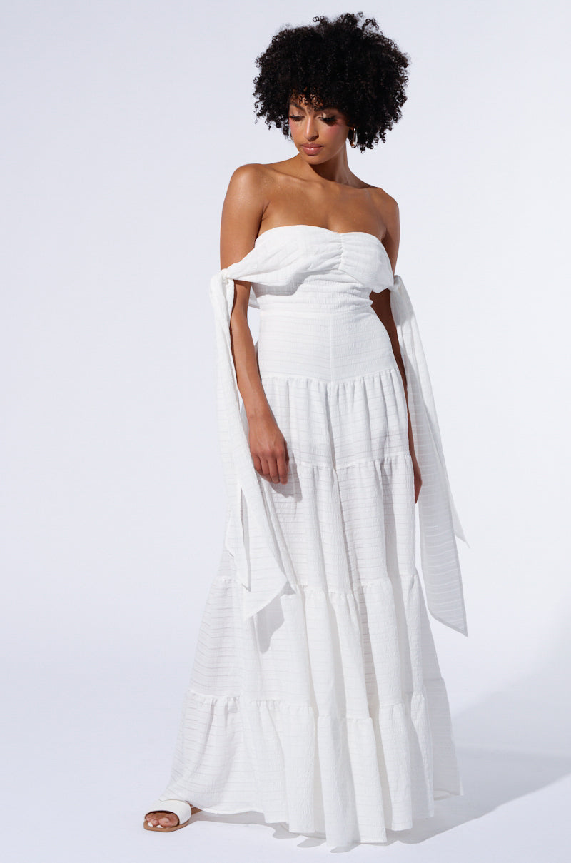 A woman is seen wearing the 'GO OFF JUMPSUIT,' characterized by its white, strapless design with off-shoulder sleeves and intricate seersucker detailing. She stands against a plain background. The jumpsuit features a ruched bodice and a tiered skirt-like bottom. The model, with curly hair, is looking down with one hand resting by her side while she wears elegant white strappy sandals.