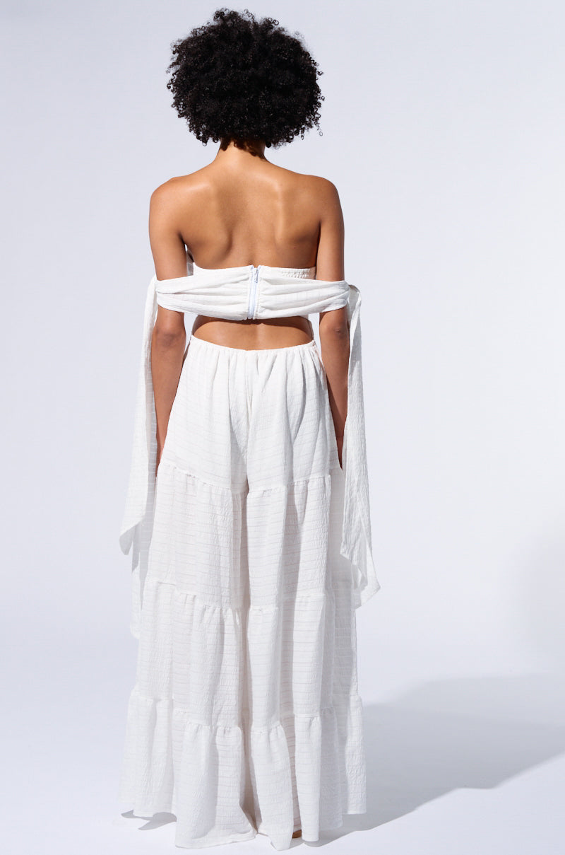 A person with curly hair stands with their back to the camera, showcasing the elegant "GO OFF JUMPSUIT." The white, strapless jumpsuit features long sleeves that hang loosely and delicate seersucker detailing. Strappy sandals complete the sophisticated look against the plain, light-colored background.