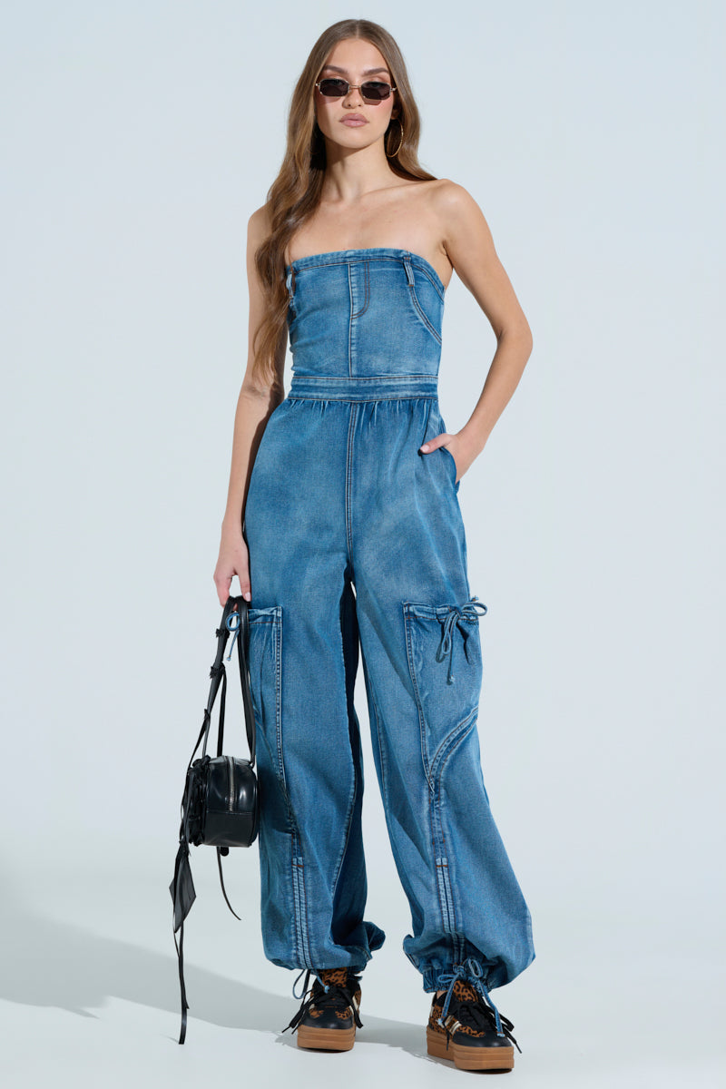 A person confidently poses in sunglasses and a JUMPING AROUND DENIM JUMPSUIT with an elasticized waist and large pockets. They hold a black bag, wear brown shoes, and stand against a plain light blue background.
