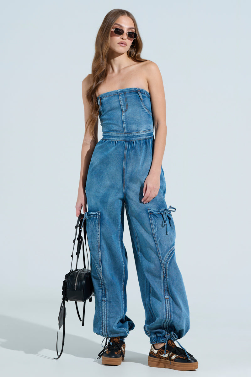 A woman in the "Jumping Around Denim Jumpsuit" with a strapless design, cargo pockets, cinched ankles, and an elastic waist holds a black handbag. Her long hair flows under sunglasses as she wears black platform sneakers against a plain background.