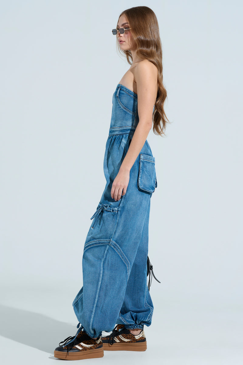 A person with long hair wears sunglasses and the JUMPING AROUND DENIM JUMPSUIT with an elasticized waist, large pockets, and tied hems, paired with brown platform sneakers against a light blue background.