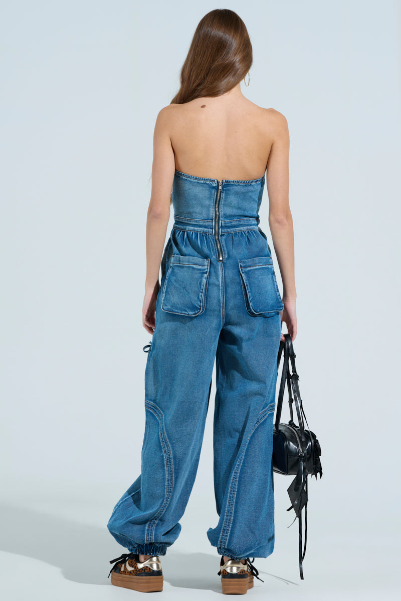 A person with long hair stands facing away, highlighting the strapless neckline and back zipper of the JUMPING AROUND DENIM JUMPSUIT. It features wide-leg pants and large pockets. They carry a tassel-accented black bag and wear brown platform shoes against a light blue background.