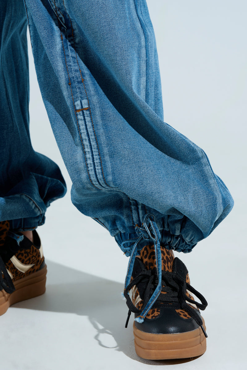 A close-up shows the JUMPING AROUND DENIM JUMPSUIT with cinched ankles, paired with black sneakers featuring a leopard print design and brown soles. The elasticized waist adds comfort and the plain white background emphasizes this casual fashion style.