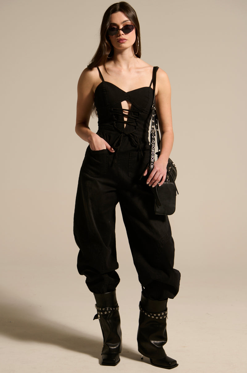 A woman confidently stands in the black "LOOKS LIKE IT JUMPSUIT," complemented by sunglasses, large hoop earrings, high studded boots, and a black bag with chain details. Her hands are tucked into the utilitarian-style pockets of her outfit.
