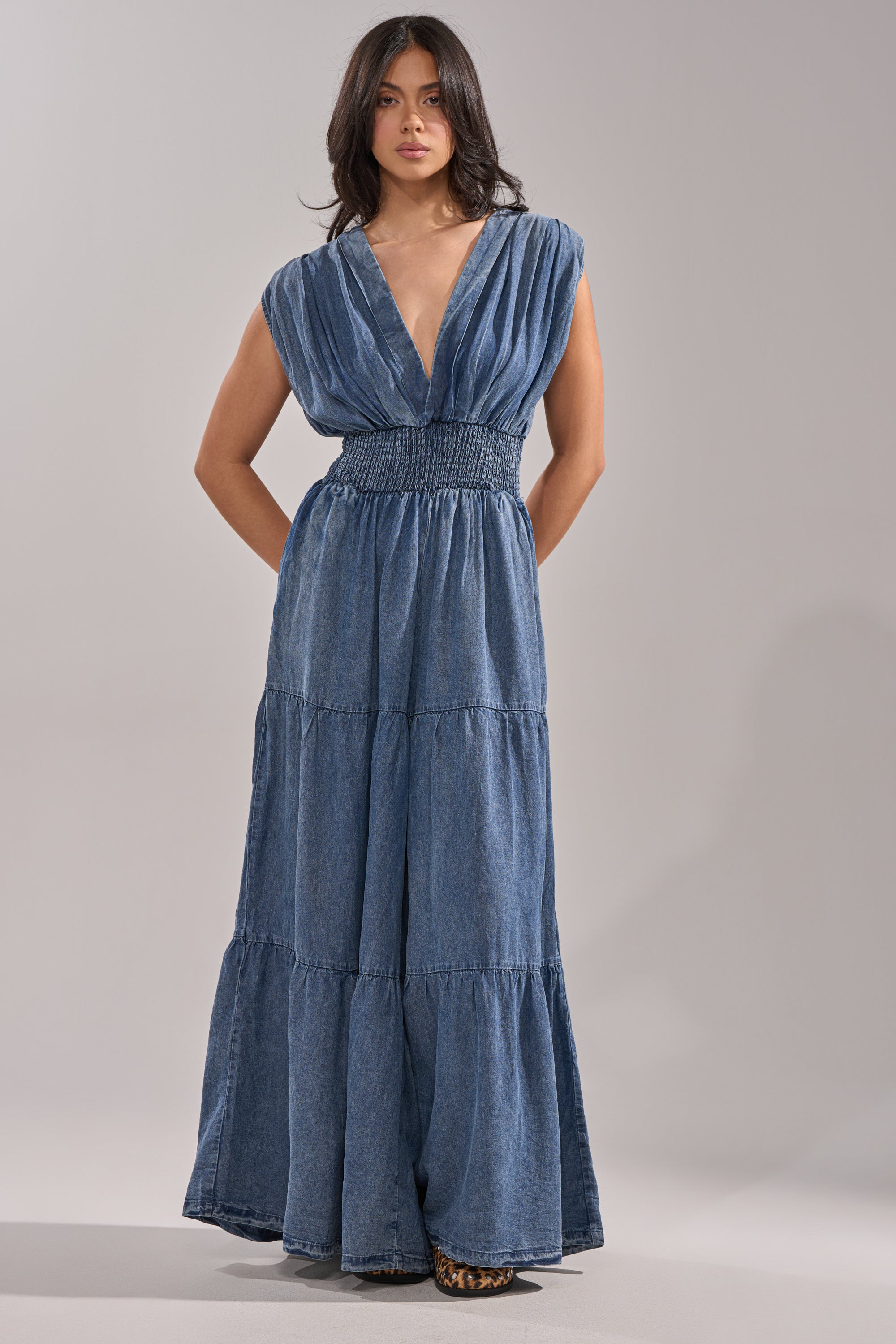 A woman poses facing forward in the KELLY DENIM JUMPSUIT, a casualwear-inspired sleeveless blue denim piece with a deep V-neck and smocked waistband. Her loose dark hair and hands behind her back create an effortlessly chic look against a neutral backdrop. 0
