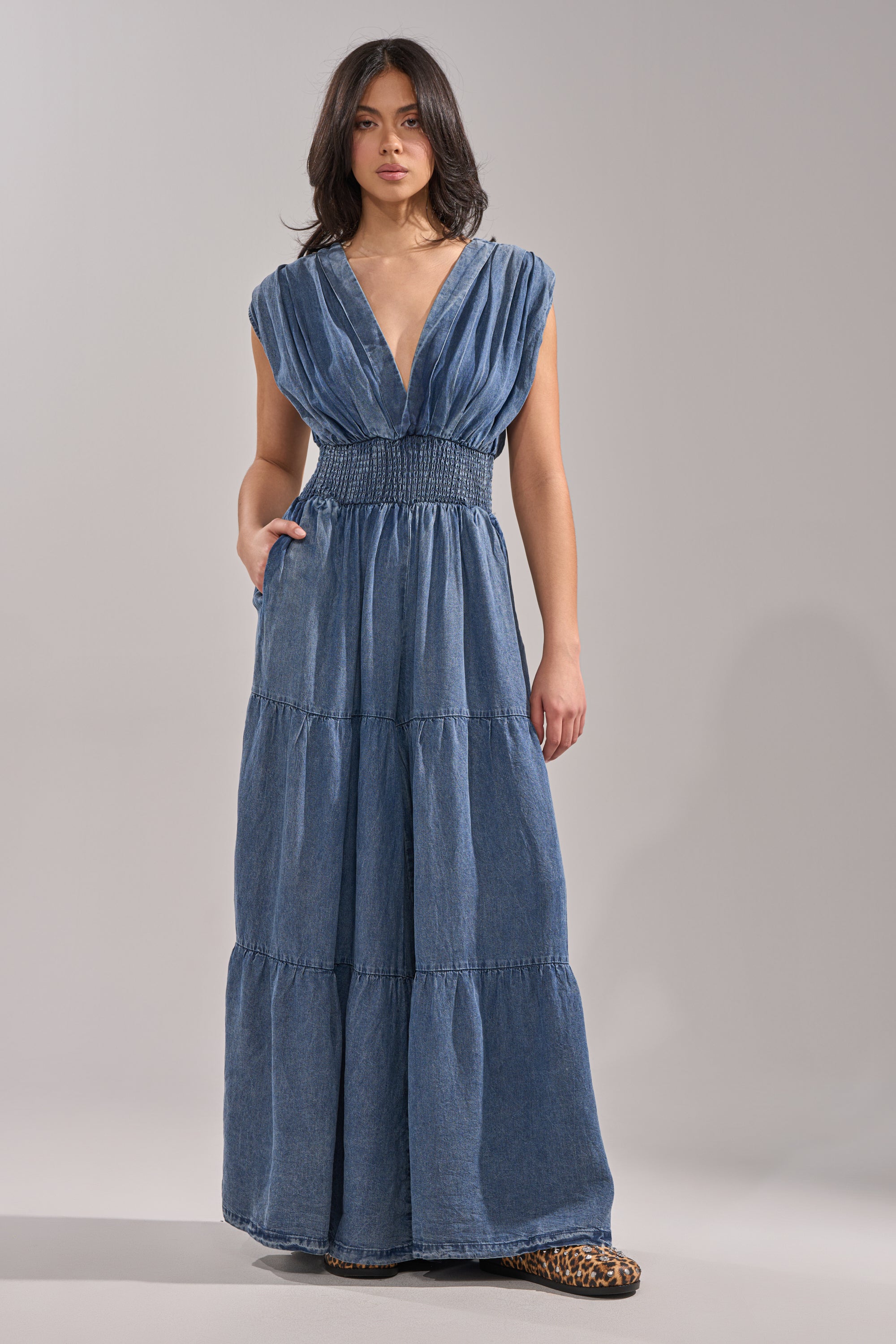 A woman models the KELLY DENIM JUMPSUIT—a sleeveless, deep V-neck, floor-length blue denim style with a smocked waist and tiered skirt—paired with leopard print shoes, set against a light background for a chic casualwear vibe. 1