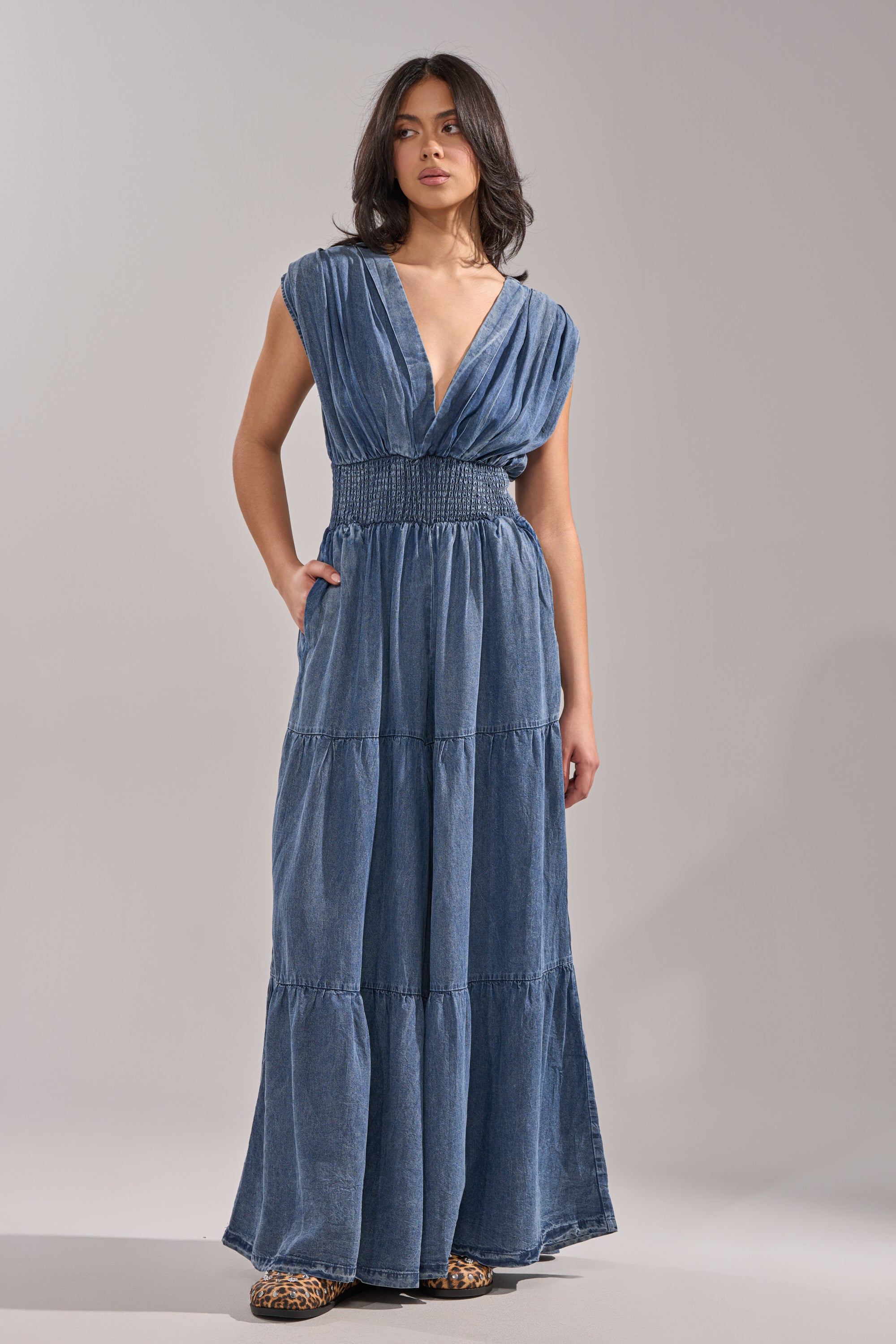 Wearing the KELLY DENIM JUMPSUIT—a sleeveless, floor-length blue denim piece with a deep V-neck and smocked waist—a woman poses casually, hands in pockets, wavy dark hair flowing, paired with leopard print shoes. 2